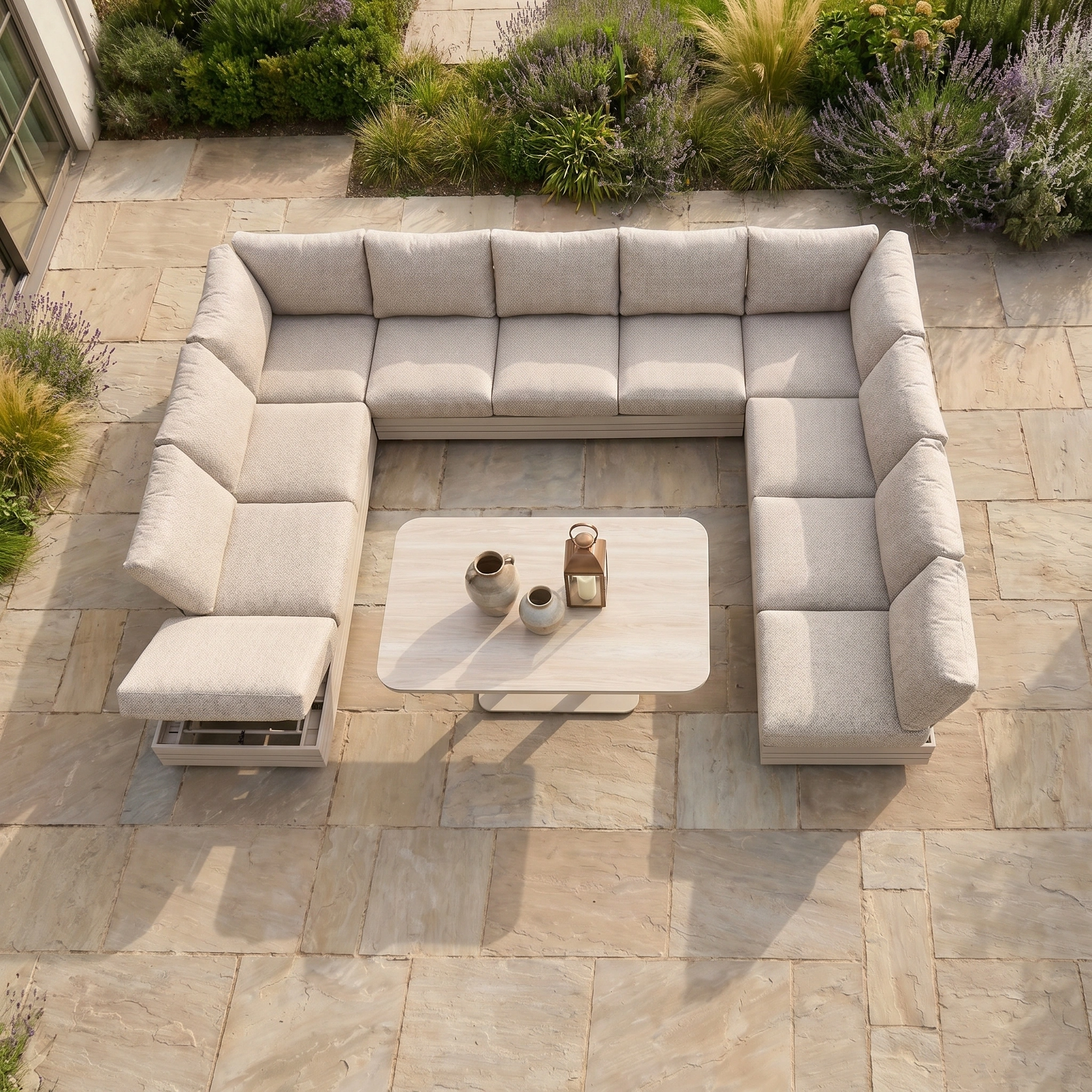The Savona Luxury Outdoor Large U-Shape Corner Sofa Set with Altura Rising Table in Oatmeal sits on a stone patio, surrounded by lush green and purple garden plants.