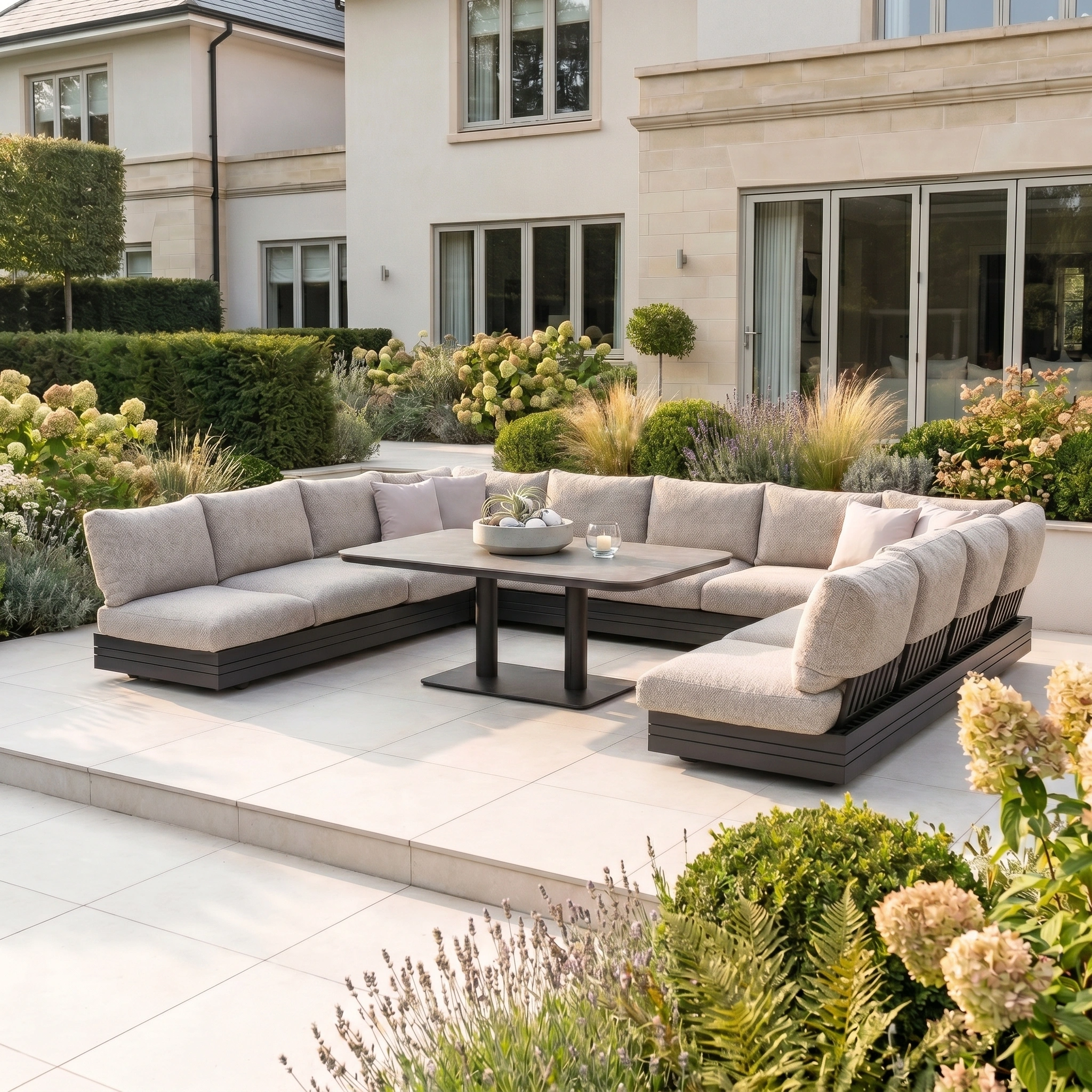 Modern patio featuring the Savona Luxury Outdoor Large U-Shape Corner Sofa Set with Altura Rising Table in Truffle, rust-proof aluminium frames, and a lush garden in front of a spacious house.