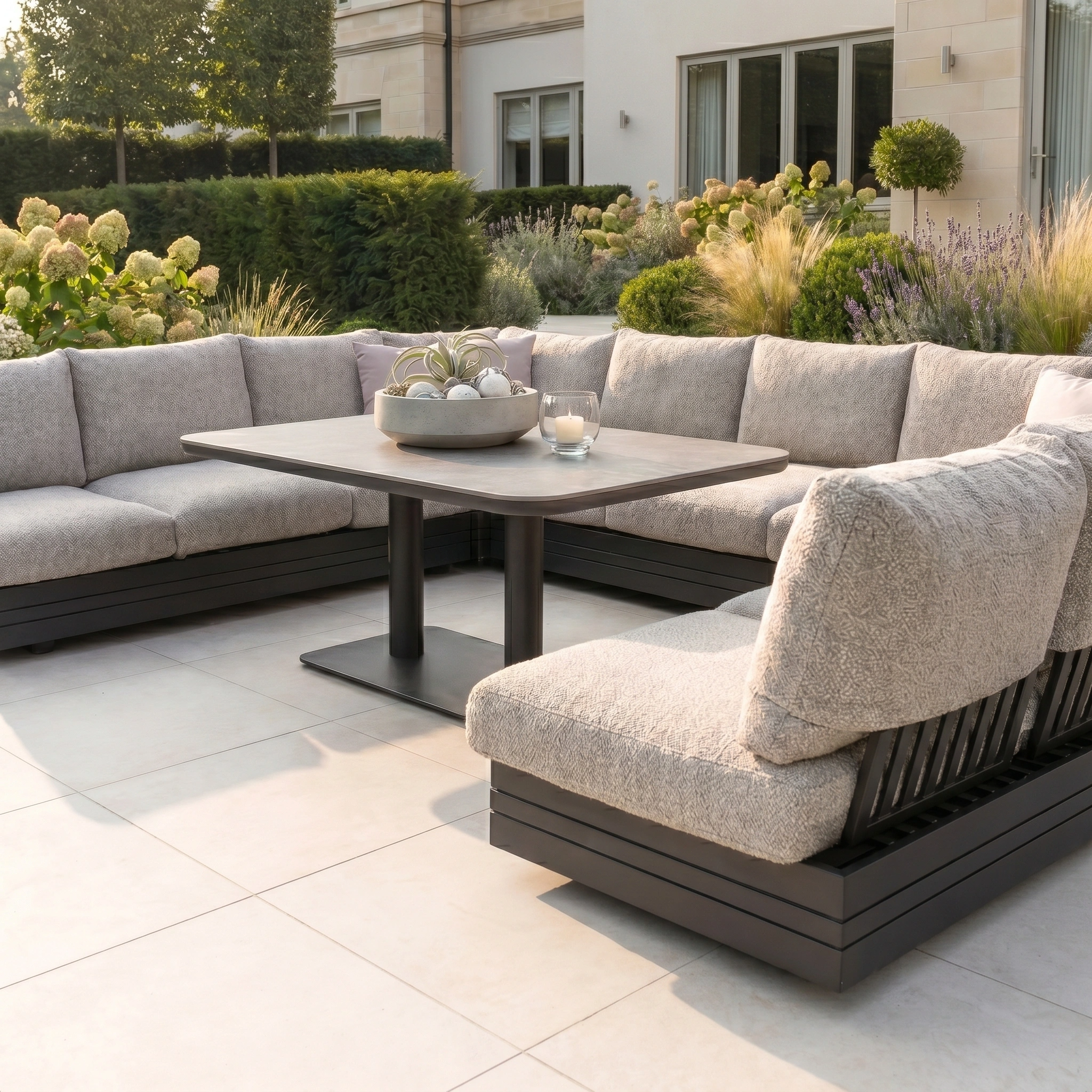 Modern patio with the Savona Luxury Outdoor Large U-Shape Corner Sofa Set and Altura Rising Table in Truffle, surrounded by lush garden landscaping.