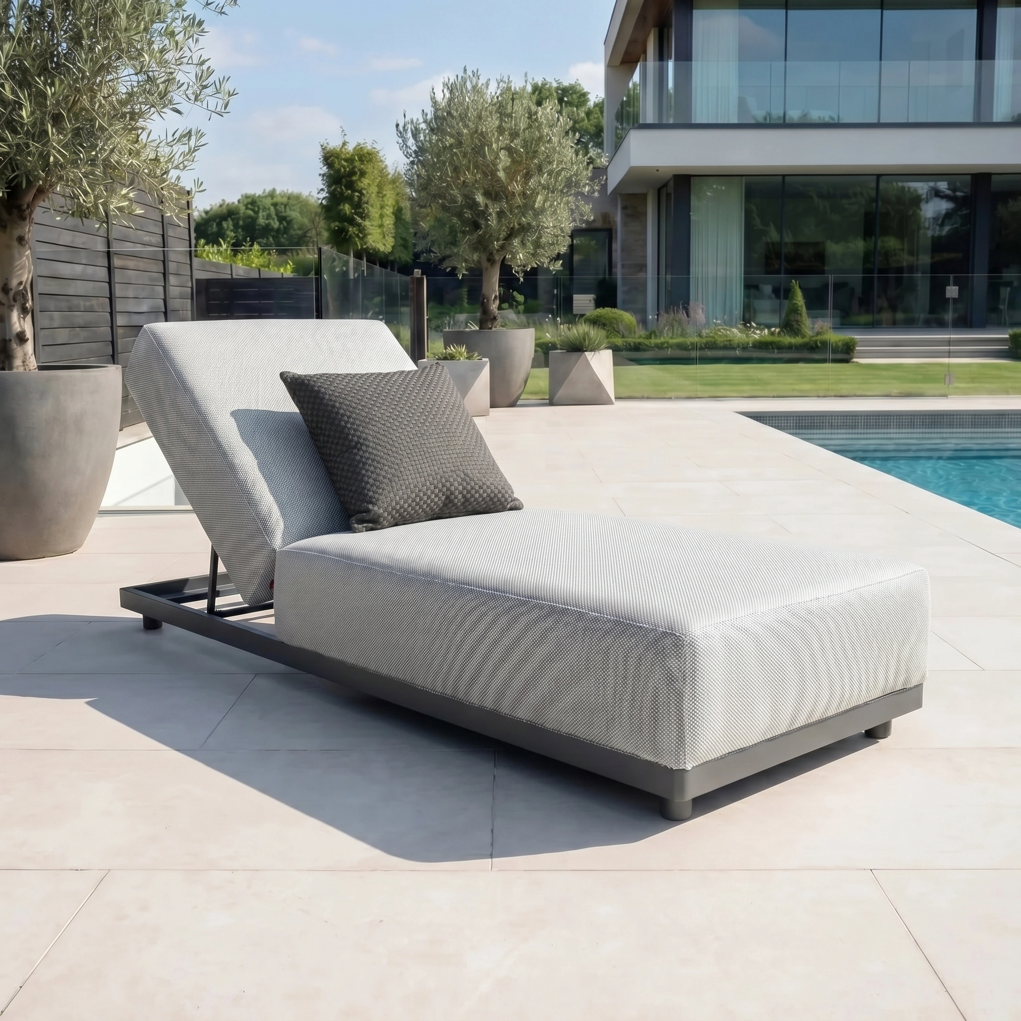 The Agora Senda Grafito Medium Scatter Cushion (45cm x 45cm), made from solution-dyed acrylic, is featured on a modern outdoor lounge chair by the pool with a contemporary house in the background.