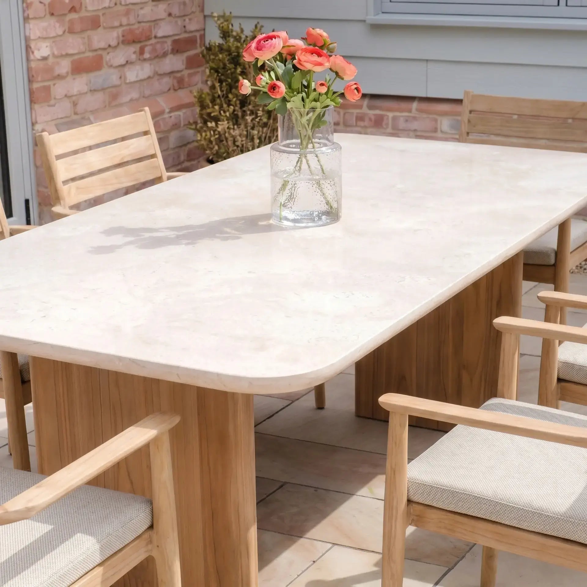 The Siena 6 Seat Dining Set in Fawn features teak dining chairs and a marble table, completed with a glass vase of pink flowers.