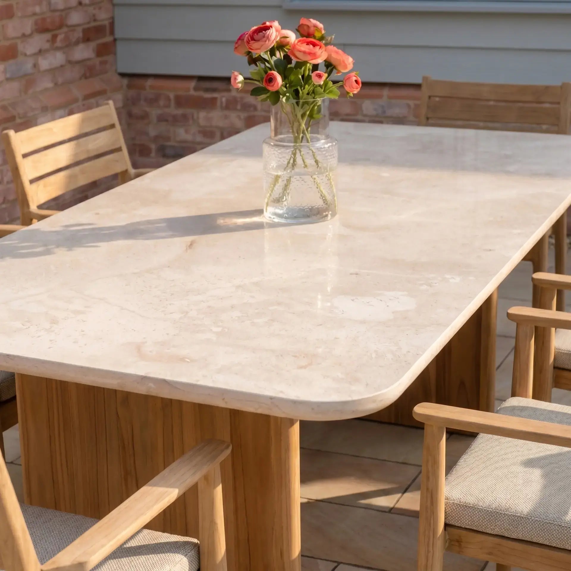 The Siena 6 Seat Dining Set in Fawn, with a vase of pink flowers on the marble table and teak chairs around it, creates an inviting outdoor patio dining space.