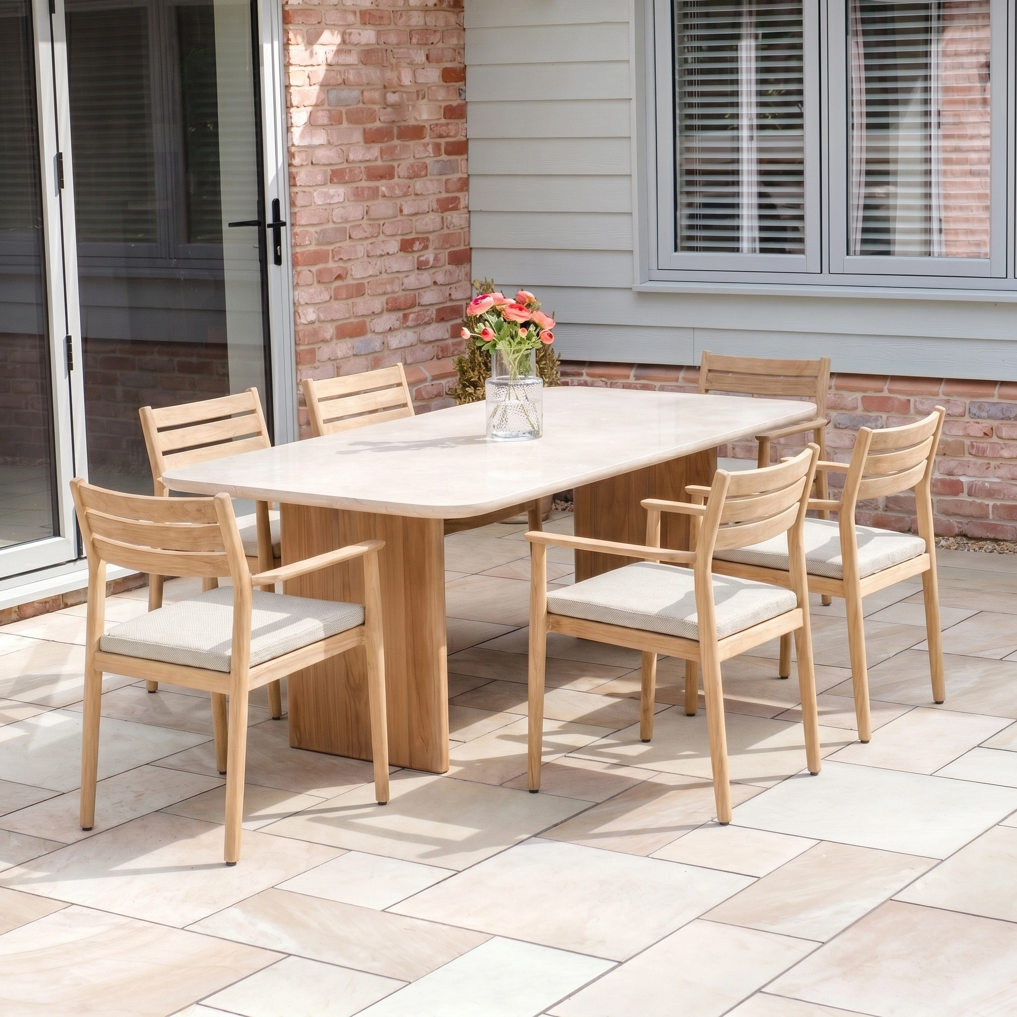The Siena 6 Seat Dining Set in Fawn features a light wooden outdoor table, six teak chairs, and a floral vase displayed on a tiled patio.