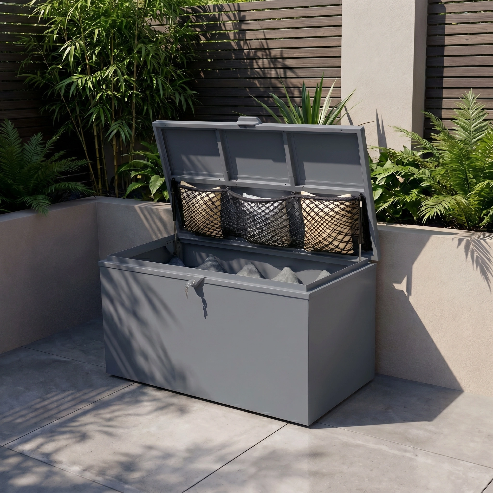 The Small Storage Box in Grey—a durable, galvanised steel storage box with an open lid displaying pillows inside—offers waterproof garden storage and sits stylishly on a patio surrounded by plants and a wooden fence.