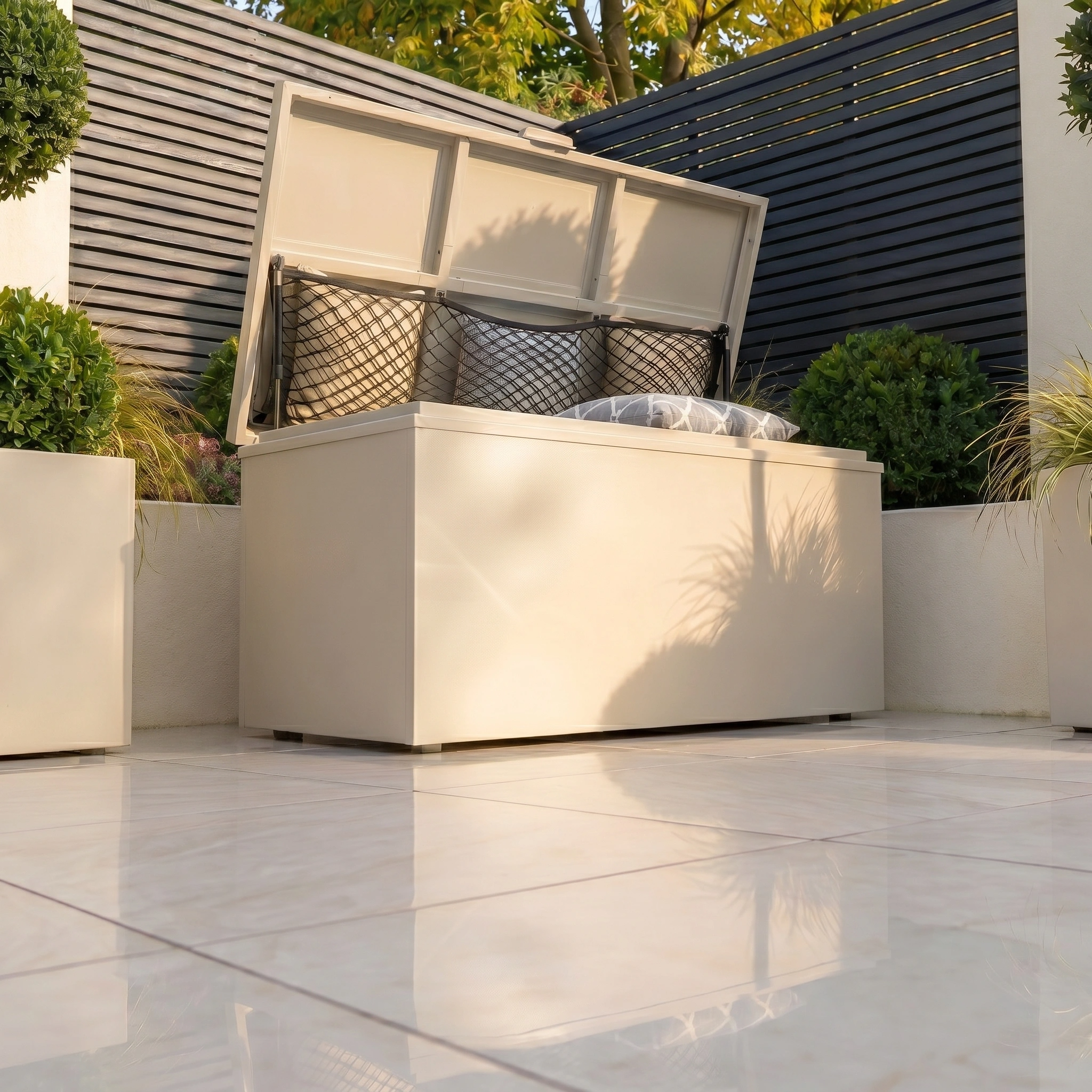The Small Storage Box in Latte sits on a tiled patio, filled with pillows and surrounded by plants and privacy fencing—a practical solution for stylish outdoor storage.