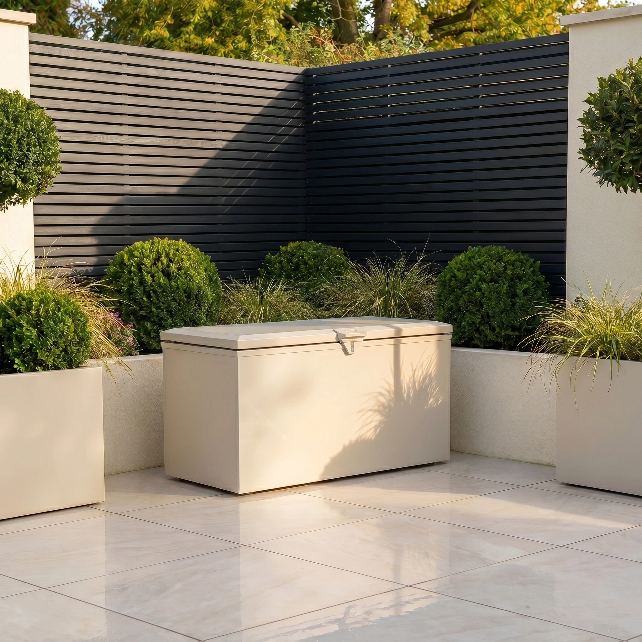 The Small Storage Box in Latte sits on a tiled patio, surrounded by potted green plants and a dark slatted fence—perfect for discreet garden storage.