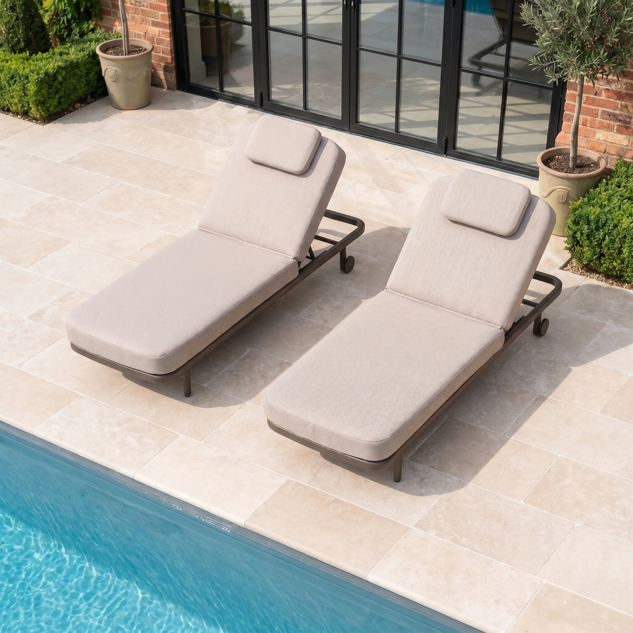 Two Sofia Sunloungers with Headrest and Wheels in Putty are placed beside a pool on a tiled patio, with potted plants nearby.