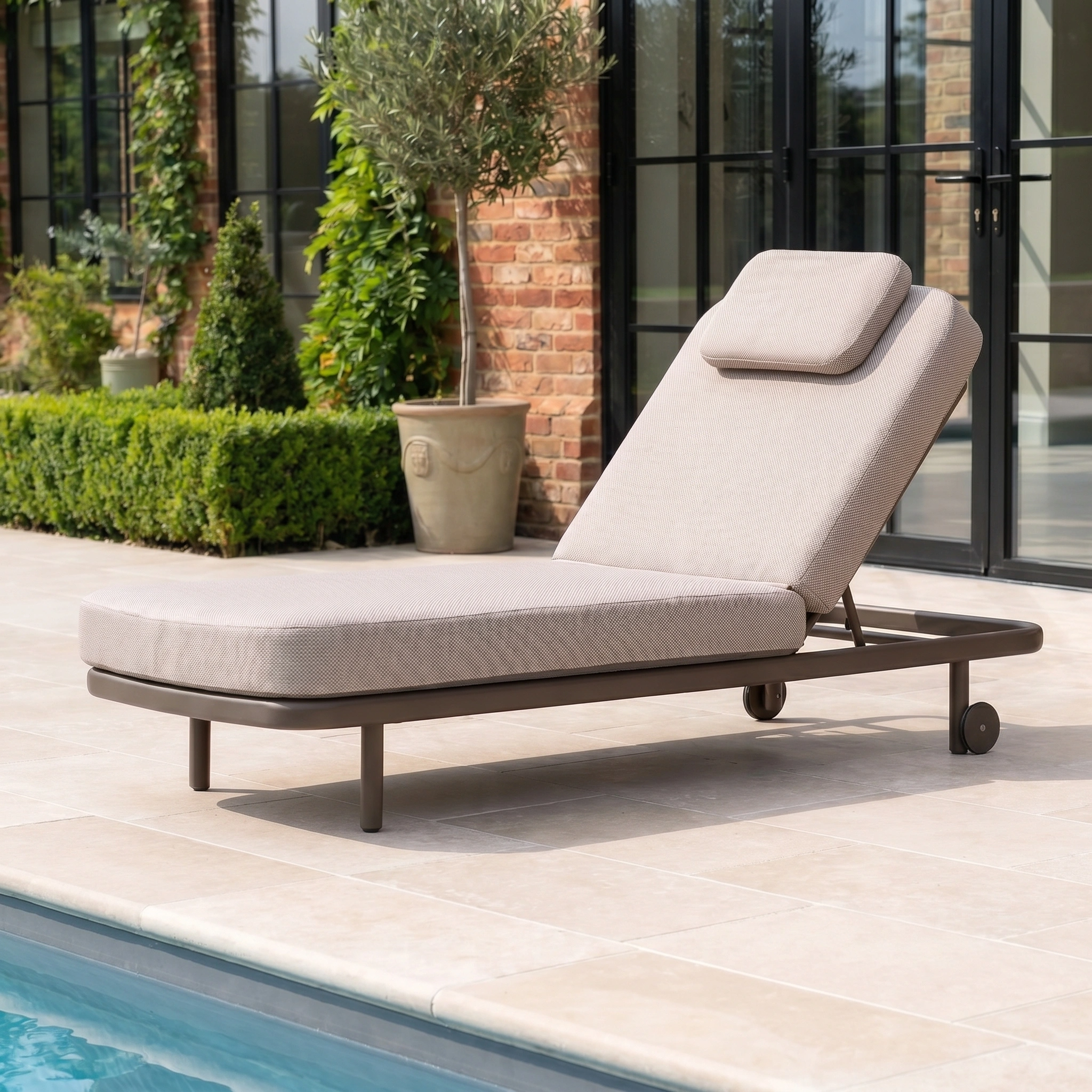 The Sofia Sunlounger with Headrest and Wheels in Putty is positioned by the poolside, featuring a cushioned design with greenery and glass doors in the background.