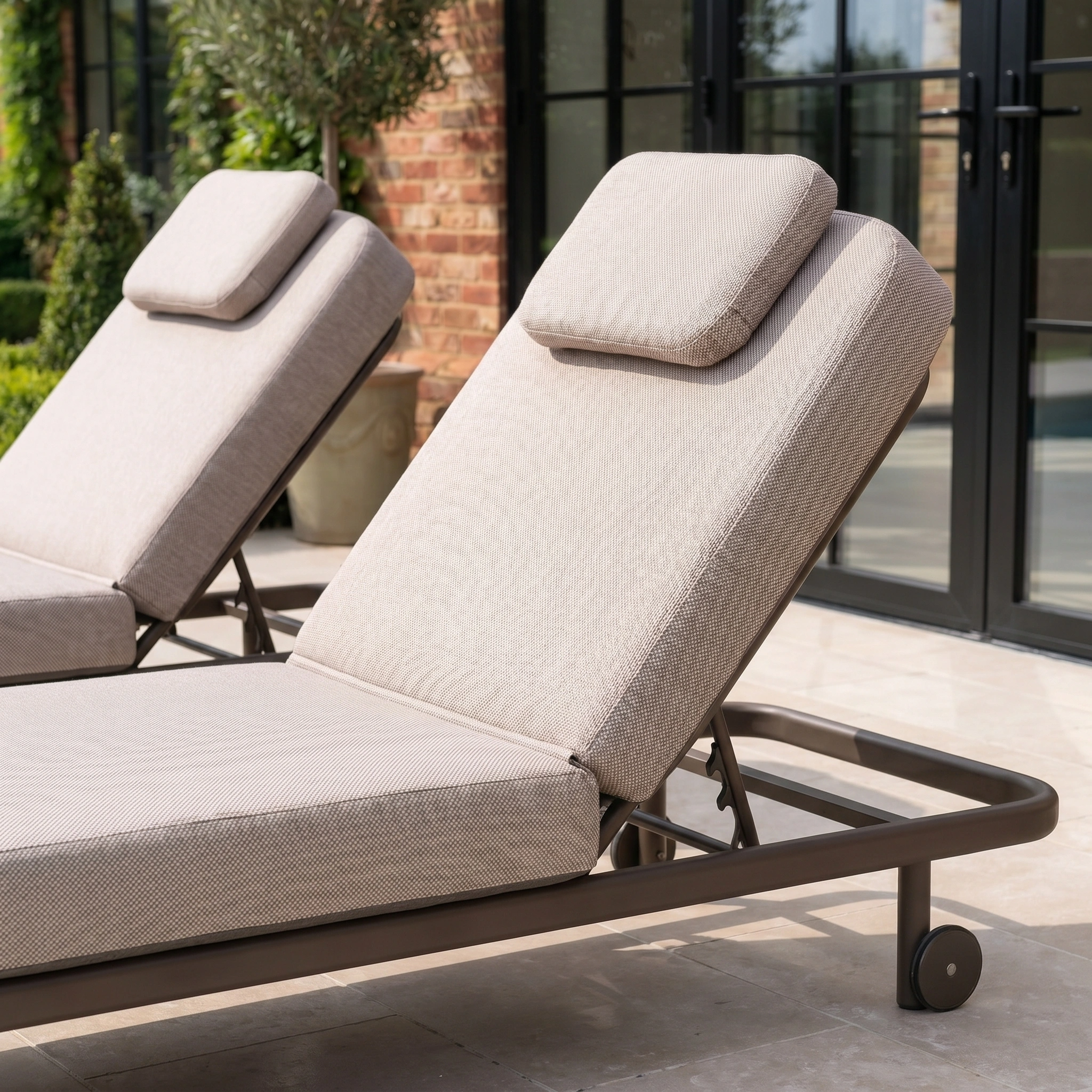 Two Sofia Sunloungers with headrests and wheels in Putty, featuring brown aluminium frames, are placed on a patio.