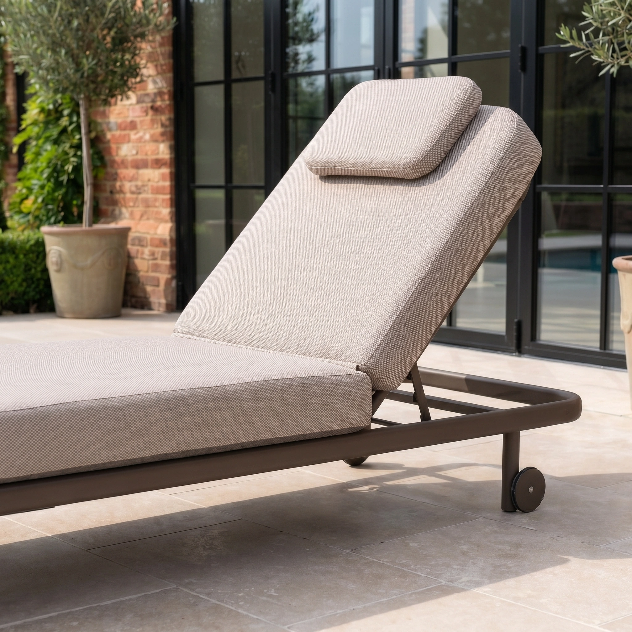 A Sofia Sunlounger with Headrest and Wheels in Putty sits on a patio by large glass doors, offering a stylish and comfortable outdoor lounging experience.