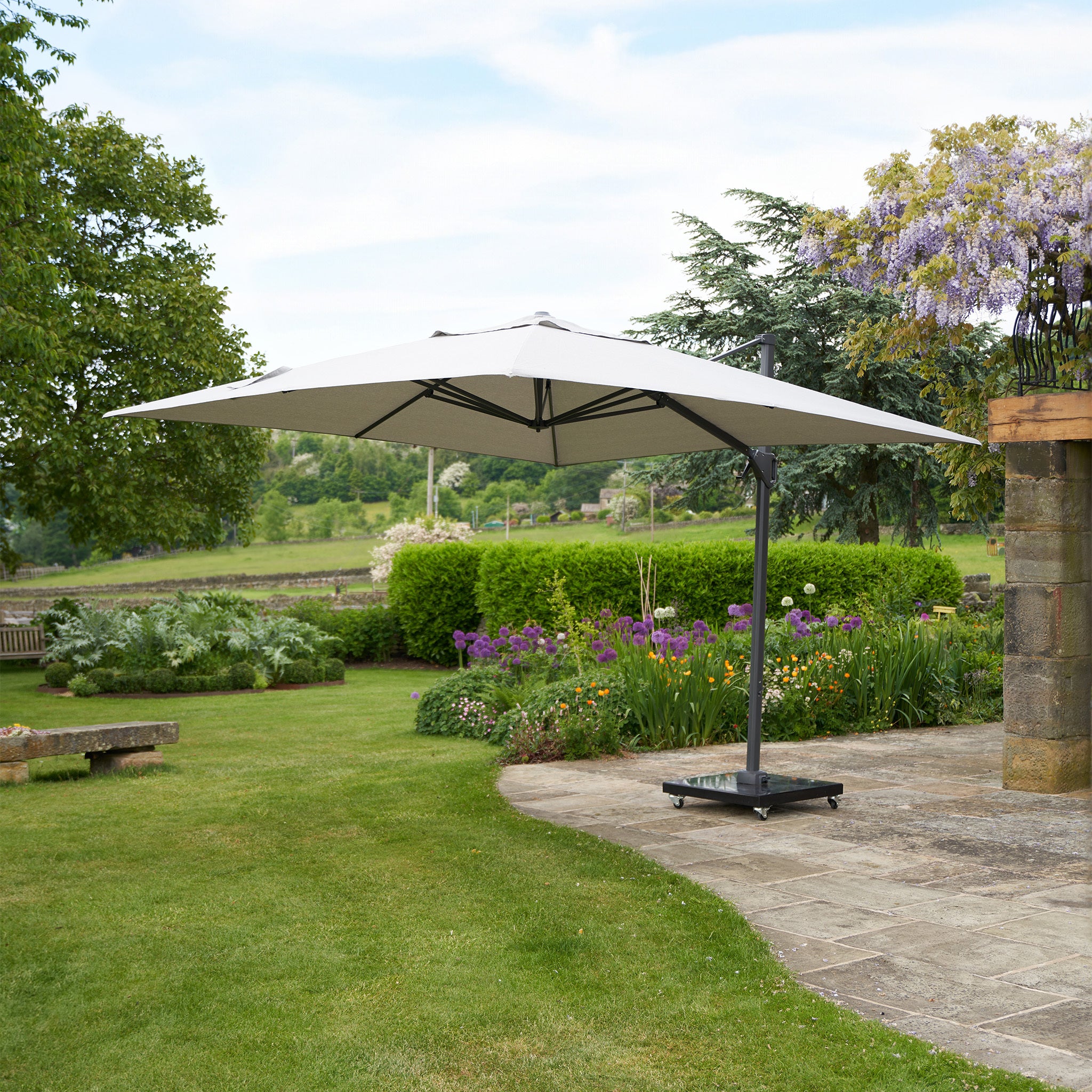 The Pacific Lifestyle Challenger T2 3m Square Cantilever Parasol in Grey features a powder-coated aluminum frame and double tilt system, providing UV protection on a garden terrace amidst greenery and flowers.