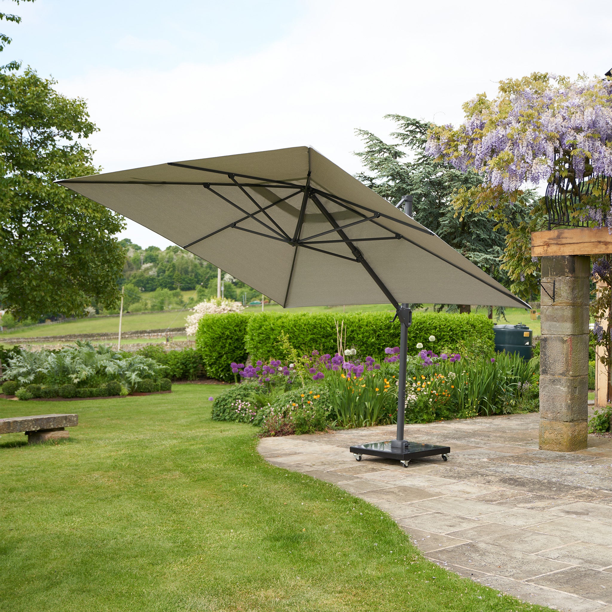 The Pacific Lifestyle - Challenger T2 3m Square Cantilever Parasol in Grey with a double tilt system and UV protection stands on a stone patio in a lush garden, surrounded by trees and flowering plants.