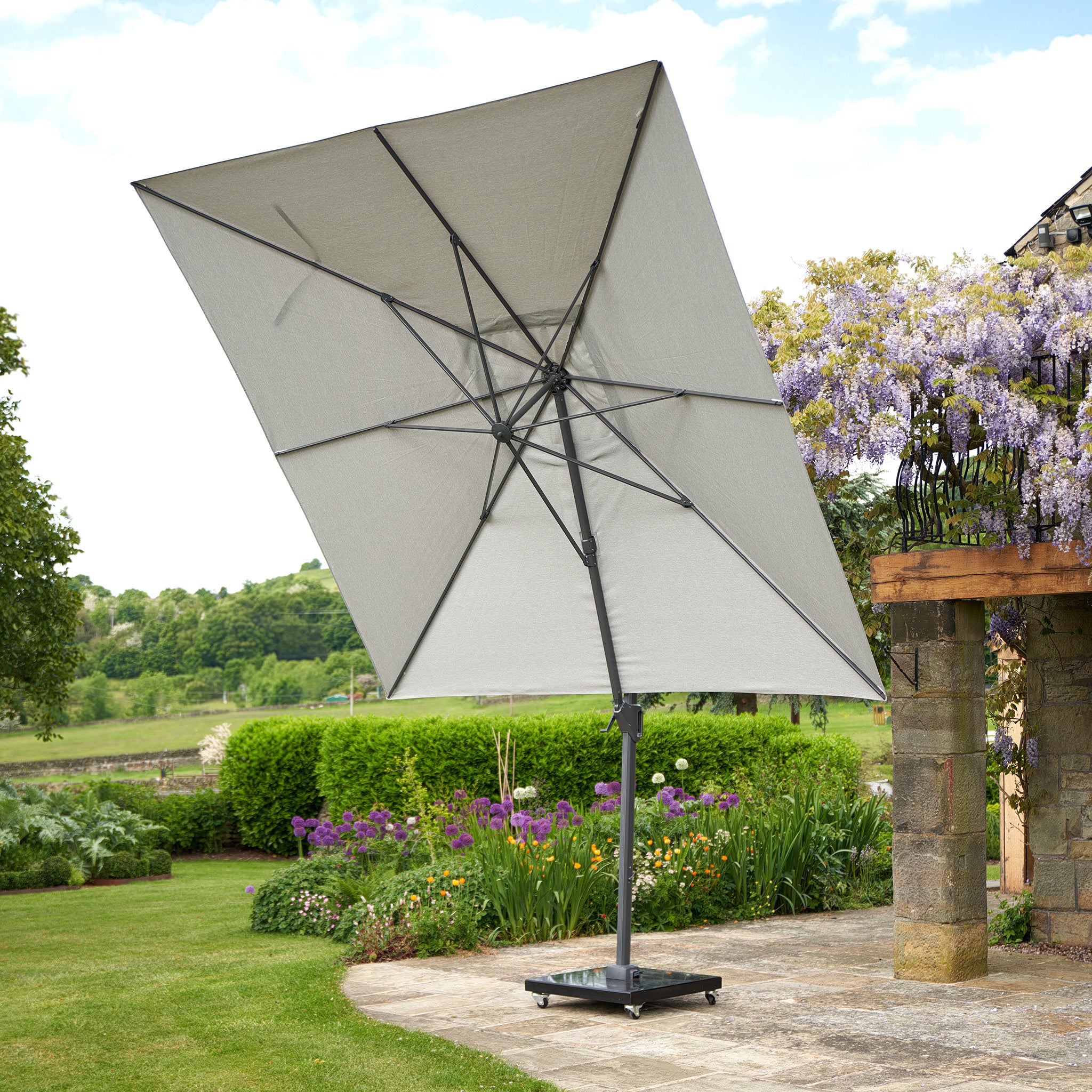 The Pacific Lifestyle Challenger T2 3m Square Cantilever Parasol in Grey stands elegantly on a stone deck, surrounded by blooming flowers and lush greenery in a garden setting.