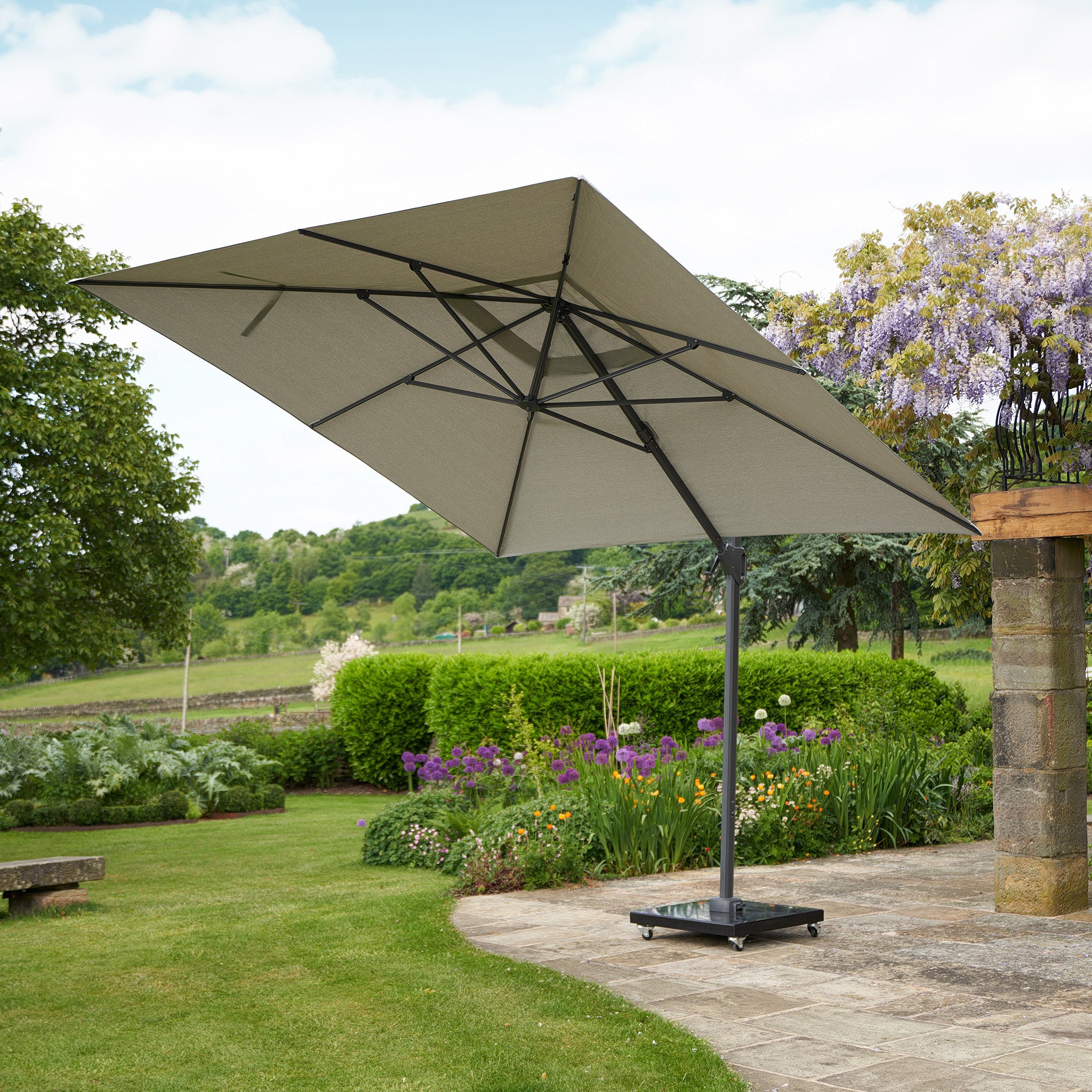 The Challenger T2 3m Square Cantilever Parasol in Grey from Pacific Lifestyle stands on a patio amid lush greenery and blooming flowers, boasting a double tilt system and a powder-coated aluminum frame for durability and UV protection.