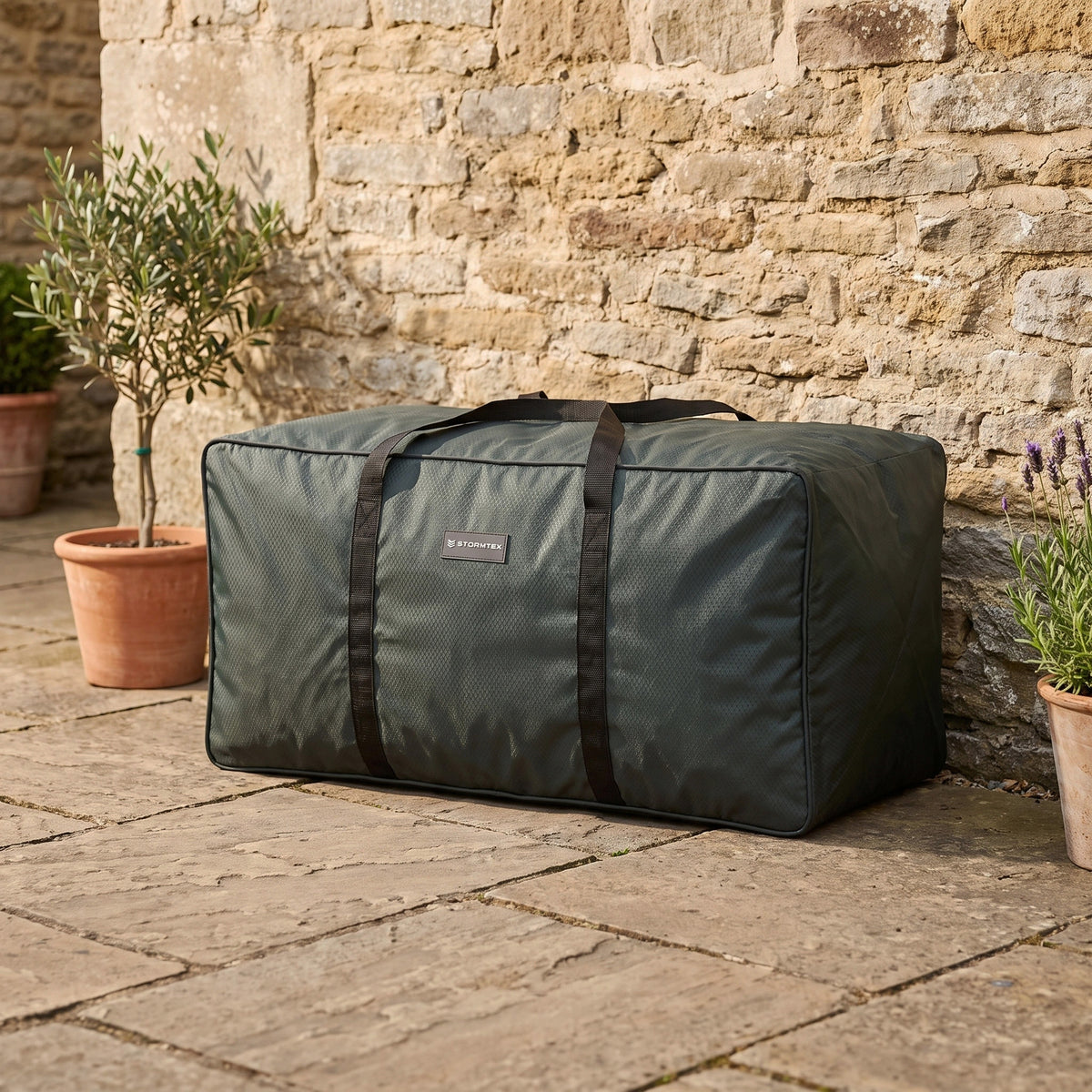 The Stormtex Cushion Storage Bag (168 x 82 x 95cm) in large green, featuring black handles, sits on a stone patio near potted plants and a stone wall, offering practical and stylish storage for your cushions.