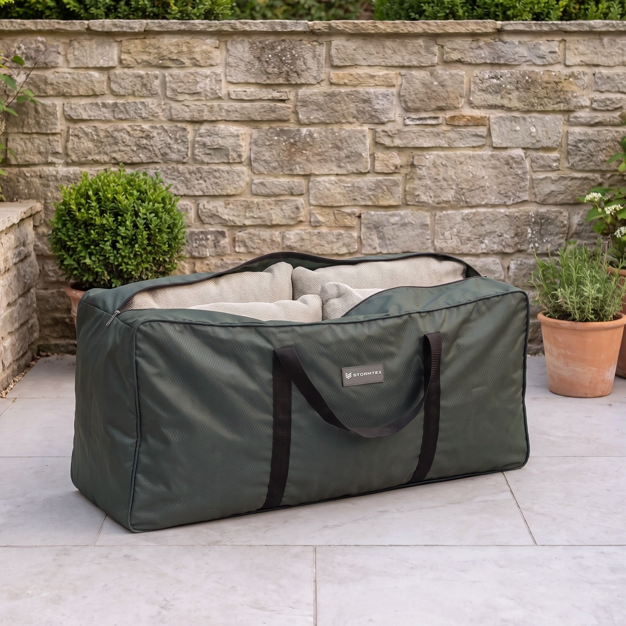 The Stormtex Cushion Storage Bag 168 x 82 x 95cm, featuring black handles and a spacious green design, sits on a patio near potted plants and a stone wall, offering practical and stylish storage for cushions.