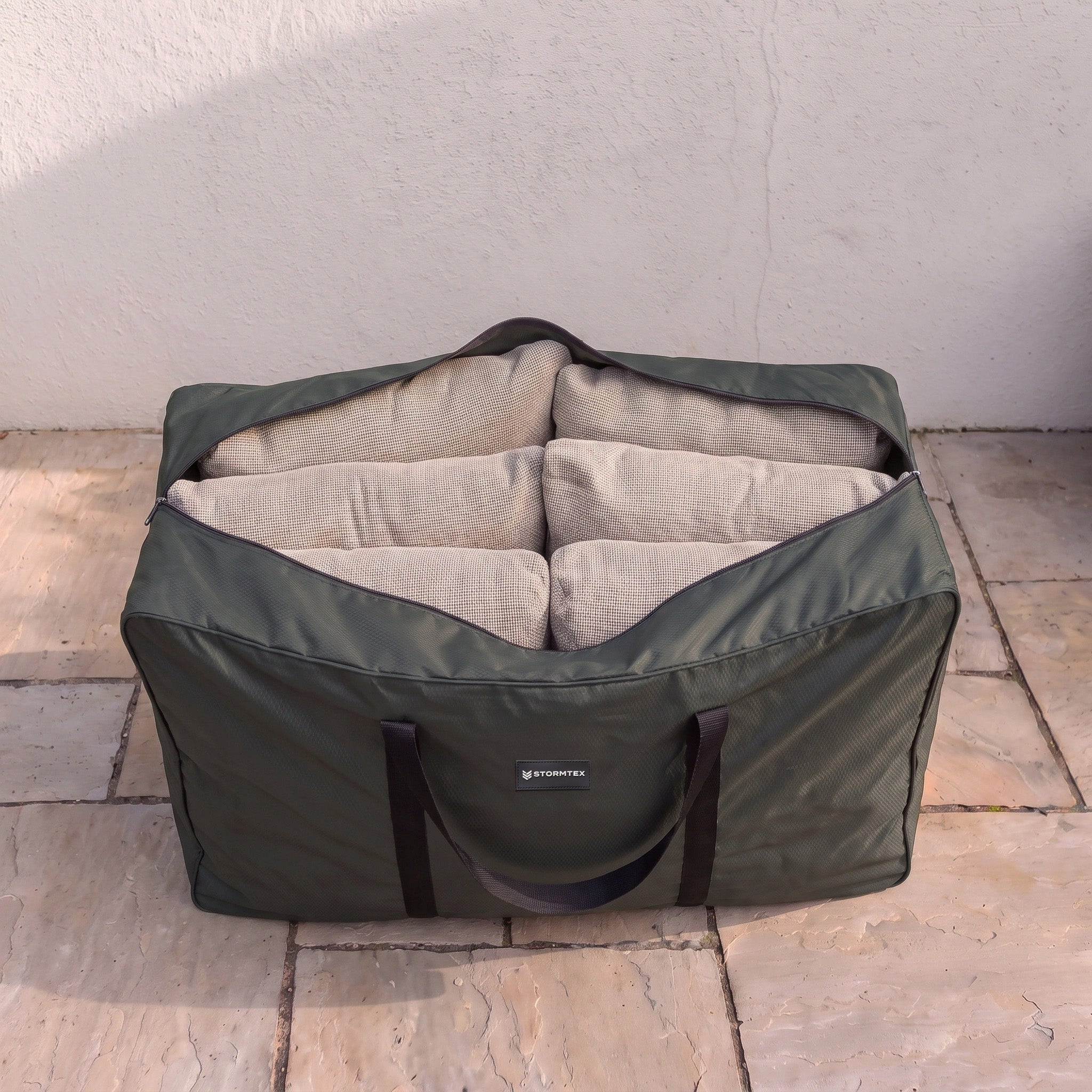 The Stormtex Cushion Storage Bag (145 x 100 x 100cm) in green, with black handles, securely holds four beige cushions on a tiled outdoor floor. Its waterproof Stormtex cover ensures your cushions remain protected from the elements.
