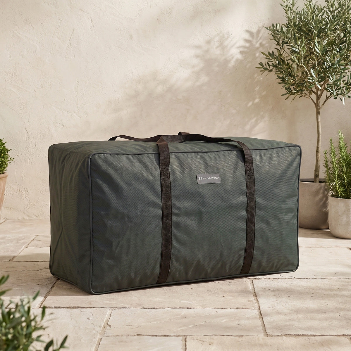 A Stormtex Cushion Storage Bag (145 x 100 x 100cm) in green with black straps is placed on a tiled patio beside potted plants.