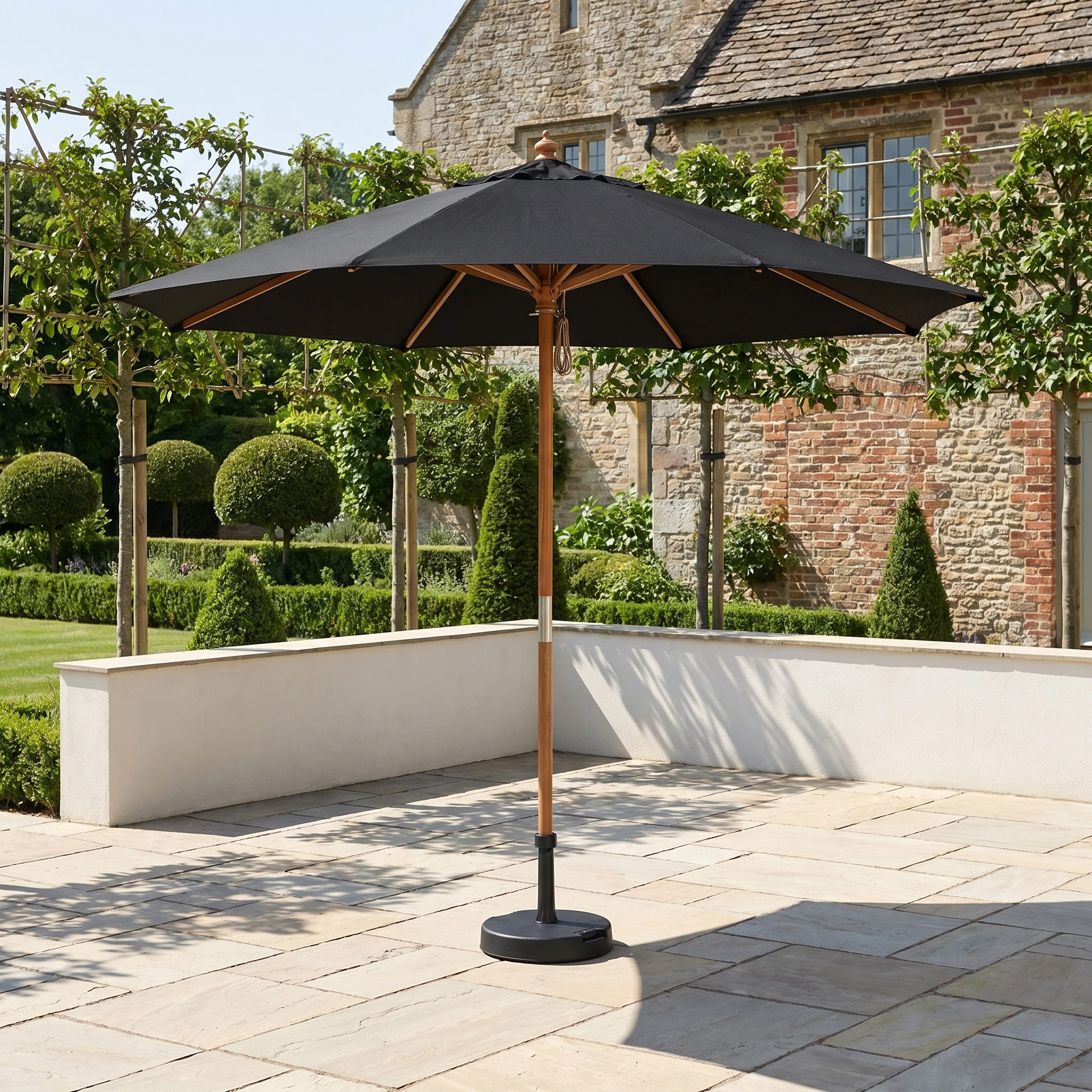 The Styx 3m Round Deluxe Wooden Parasol in Black stands on a stone terrace by a brick house, providing shade and UV protection in the landscaped garden.