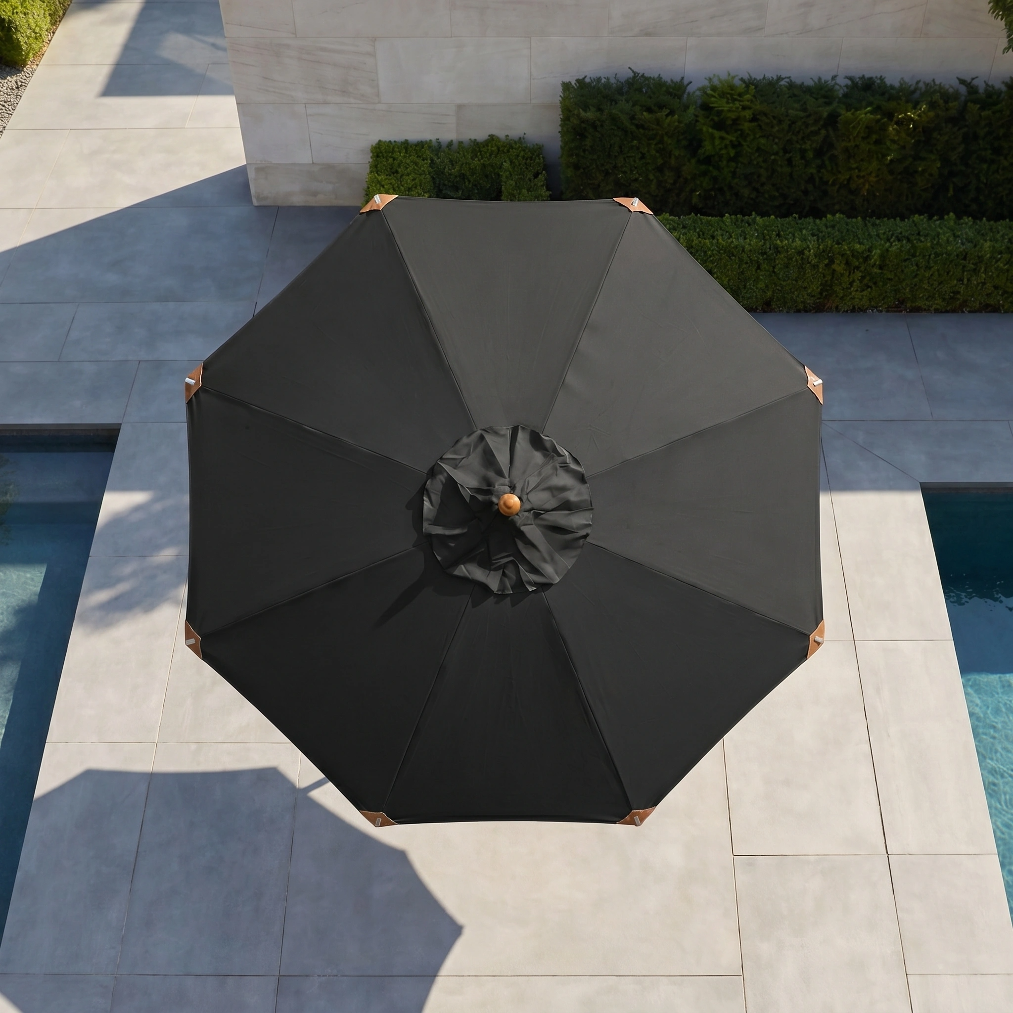 The Styx 3m Round Deluxe Wooden Parasol in Black is open over a tiled poolside area with trimmed green hedges nearby, providing stylish shade and reliable UV protection.