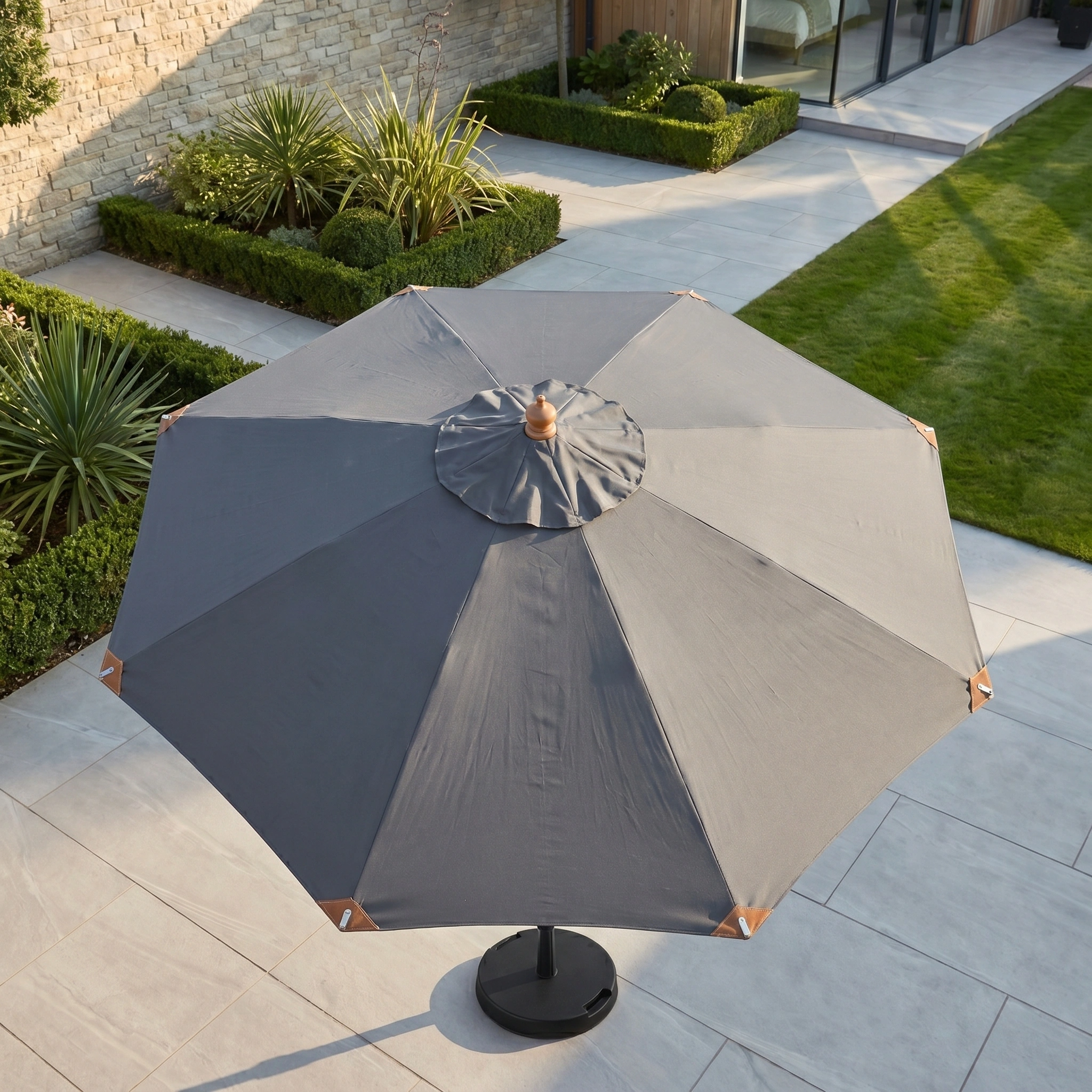 The Styx 3m Round Deluxe Wooden Parasol in Grey sits on a tiled terrace, overlooking lush green grass and a landscaped garden.