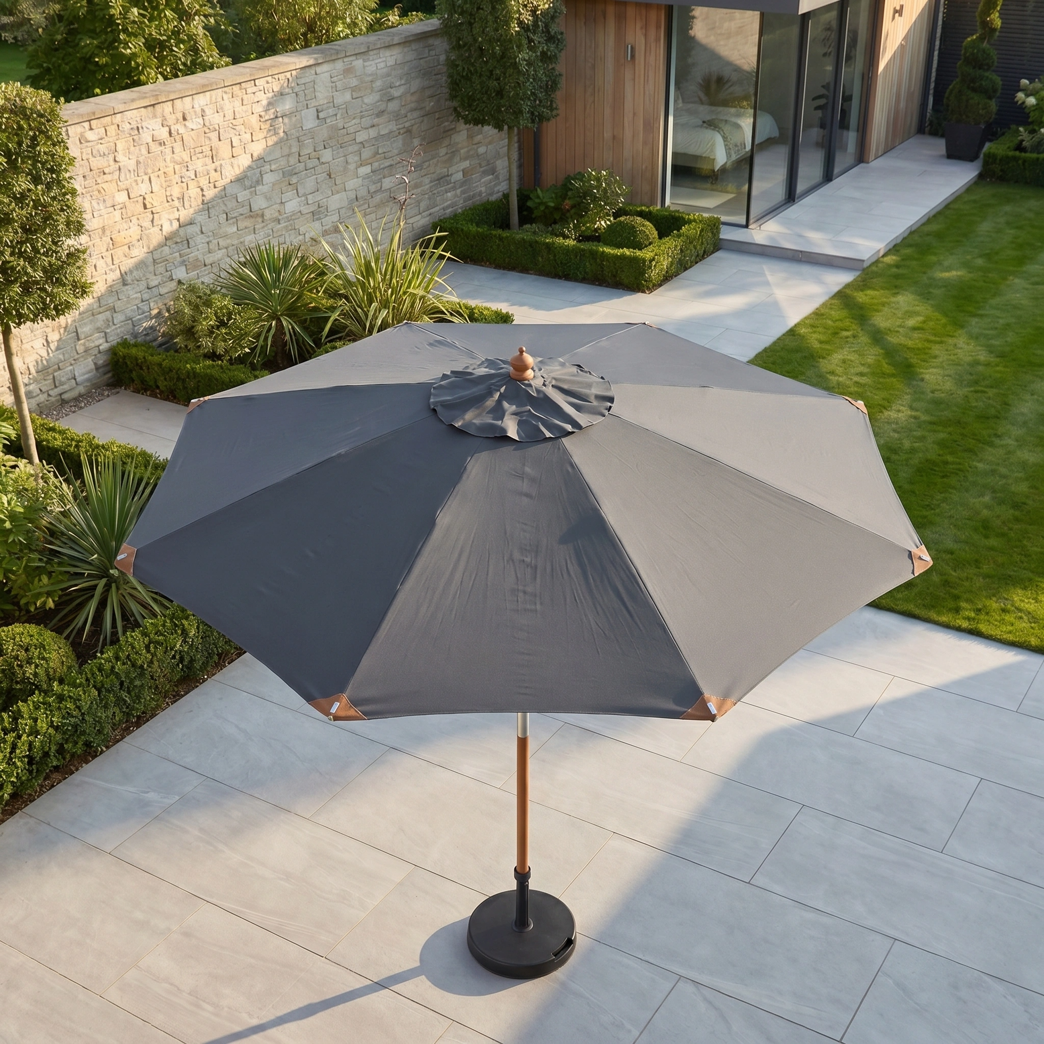 The Styx 3m Round Deluxe Wooden Parasol in Grey stands on a tiled terrace next to a modern house, overlooking a garden with neatly trimmed hedges.