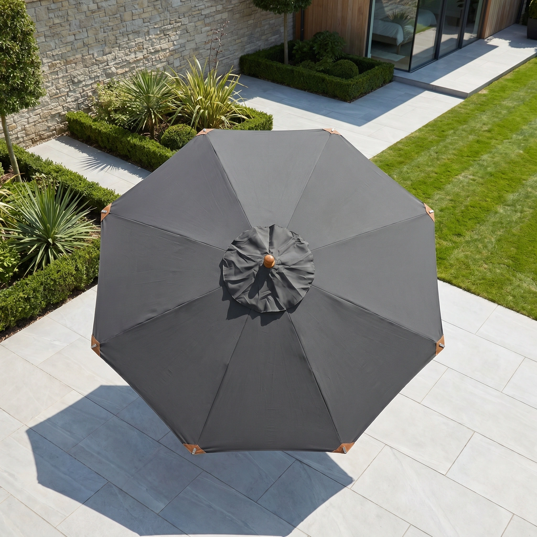 The Styx 3m Round Deluxe Wooden Parasol in Grey is shown from above on a stone patio next to neatly trimmed grass and landscaped plants.