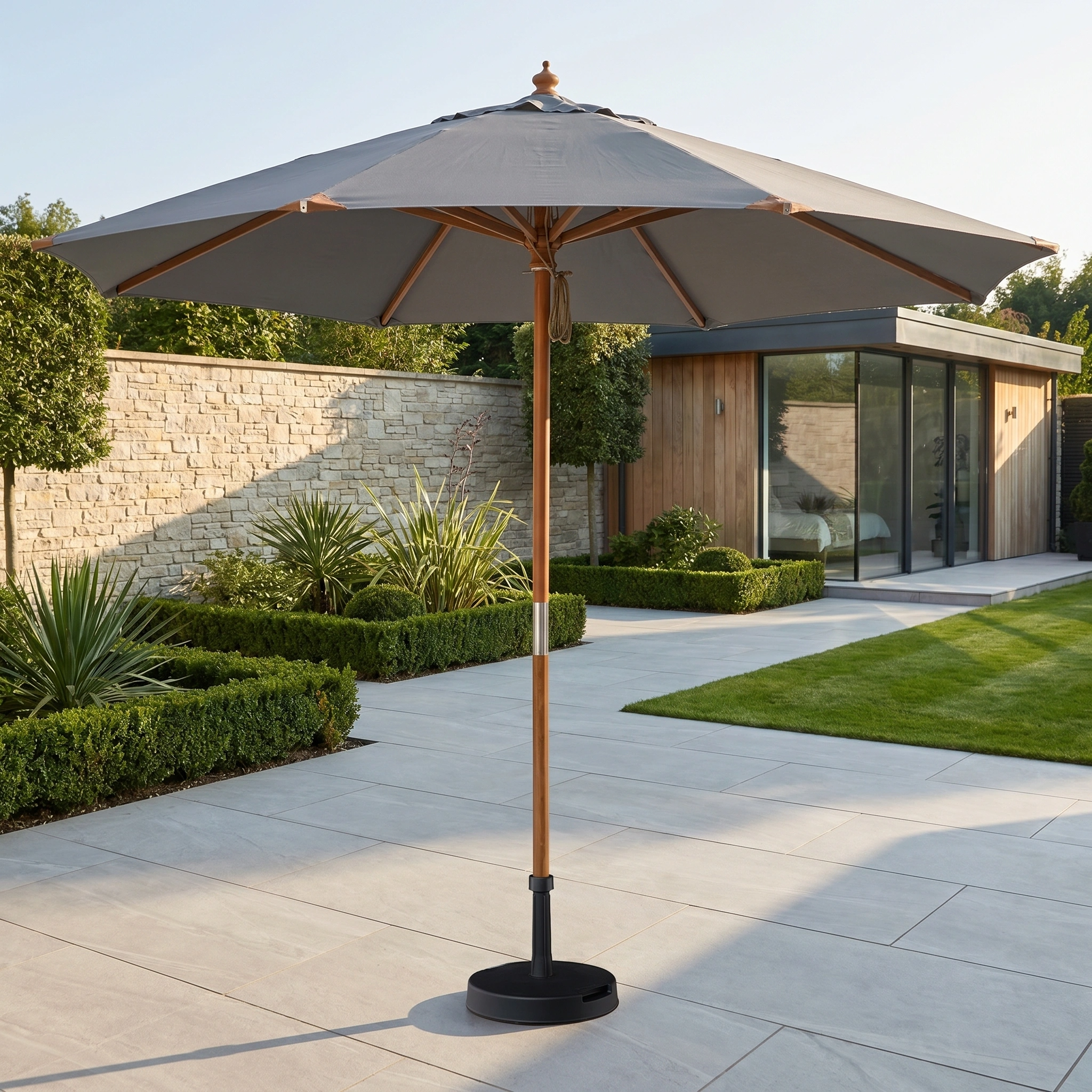The Styx 3m Round Deluxe Wooden Parasol in Grey stands on a sunny patio with modern landscaping and a stylish wooden garden building.