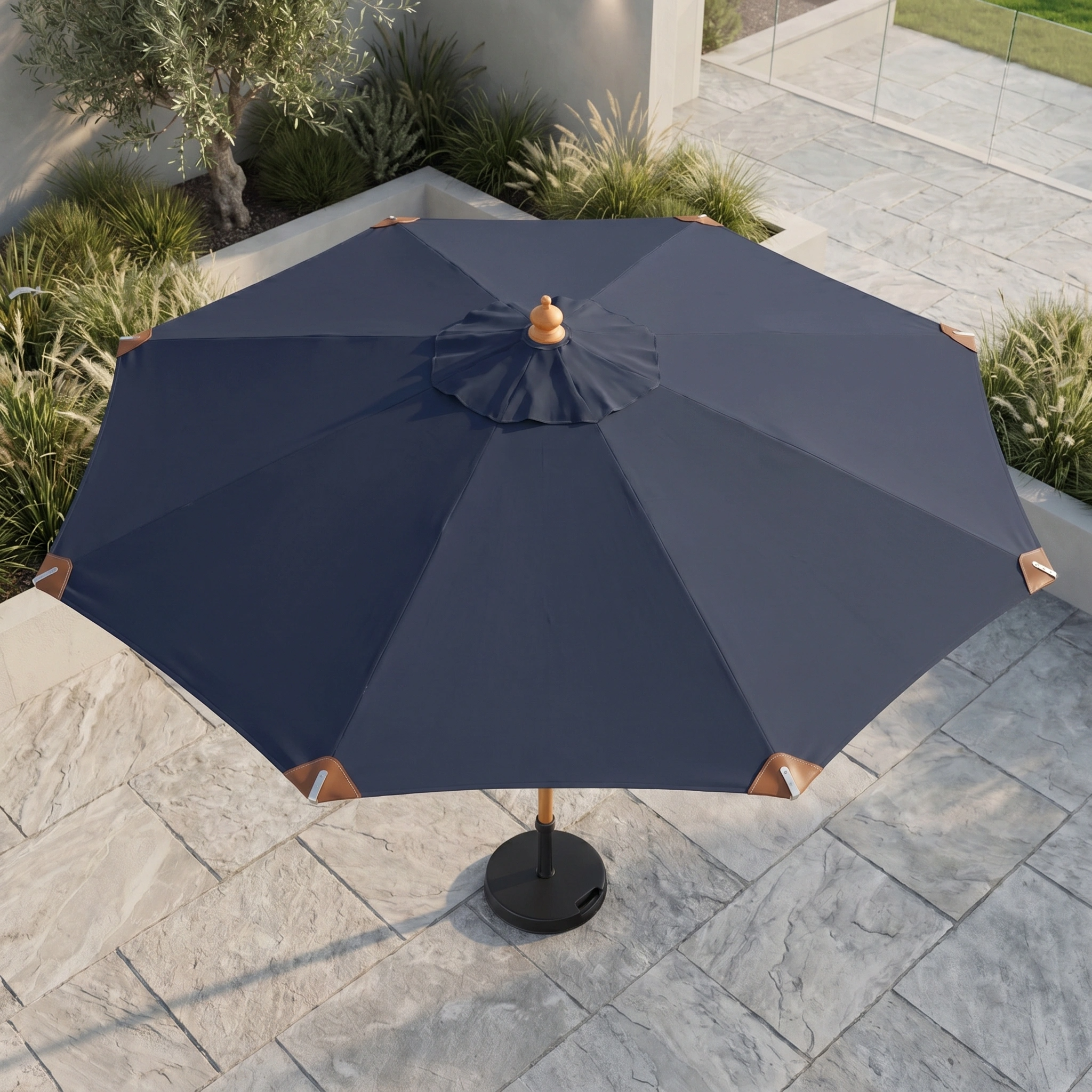 The Styx 3m Round Deluxe Wooden Parasol in Navy is open on a stone-tiled patio, surrounded by greenery and modern landscaping.