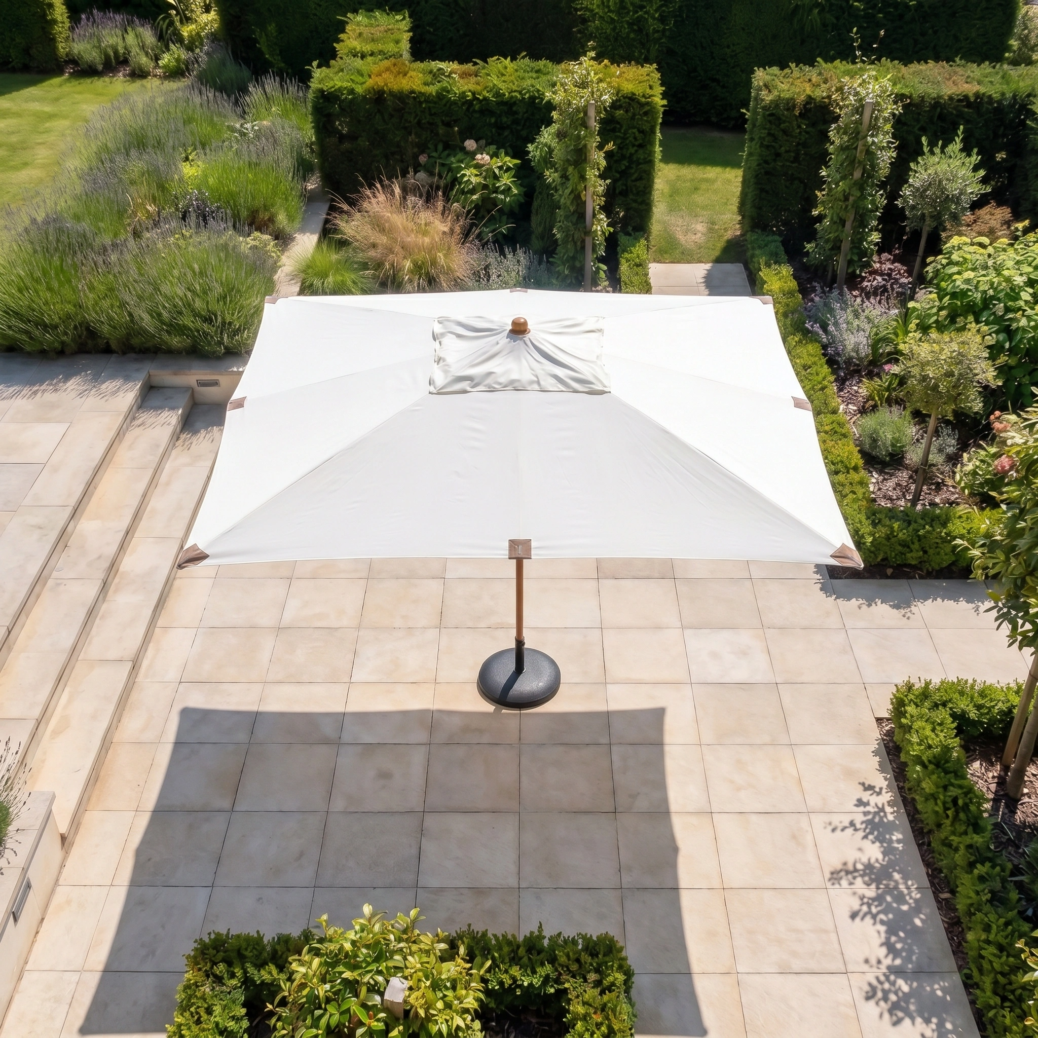The Styx 3m x 2m Rectangular Deluxe Wooden Parasol in Beige stands on a tiled terrace, framed by green hedges and lush garden landscaping.