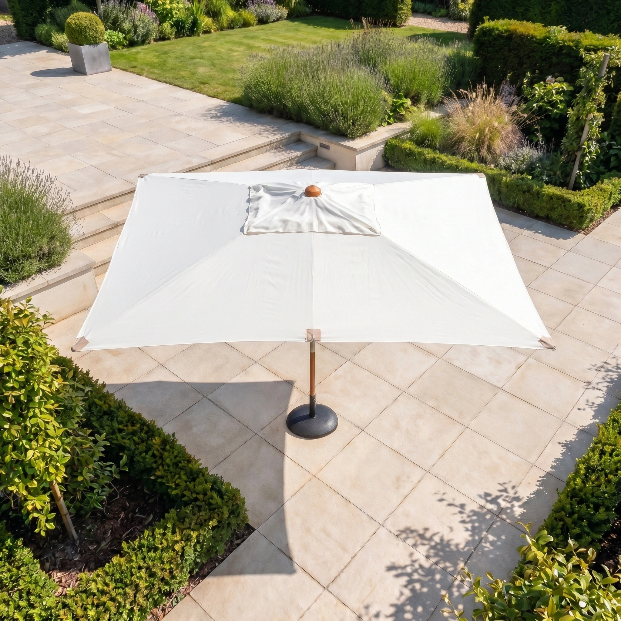 The Styx 3m x 2m Rectangular Deluxe Wooden Parasol in Beige offers shade on a tiled terrace surrounded by lush green hedges and a manicured garden.