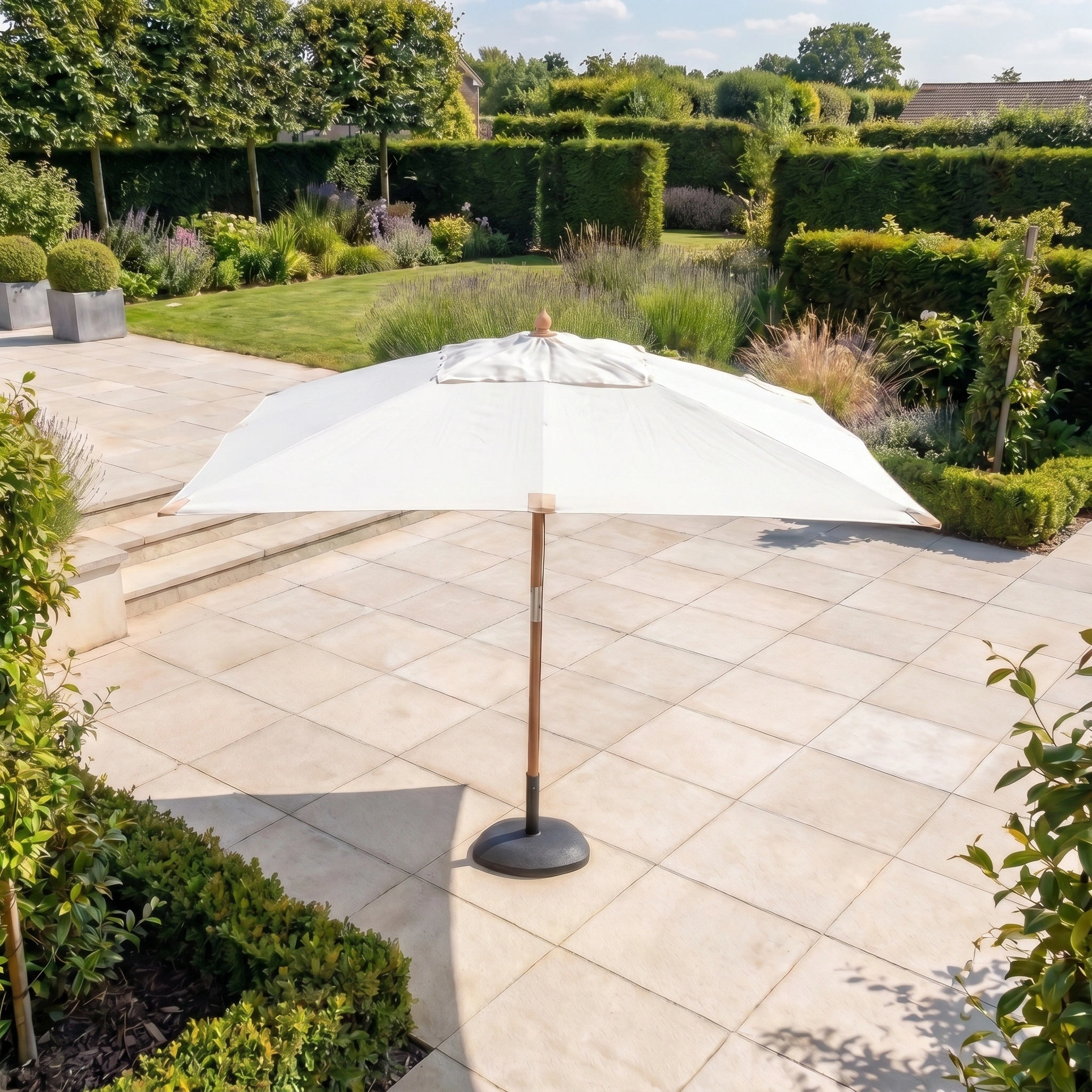 The Styx 3m x 2m Rectangular Deluxe Wooden Parasol in Beige stands on a tiled terrace amid a landscaped garden with lush greenery and shrubs.