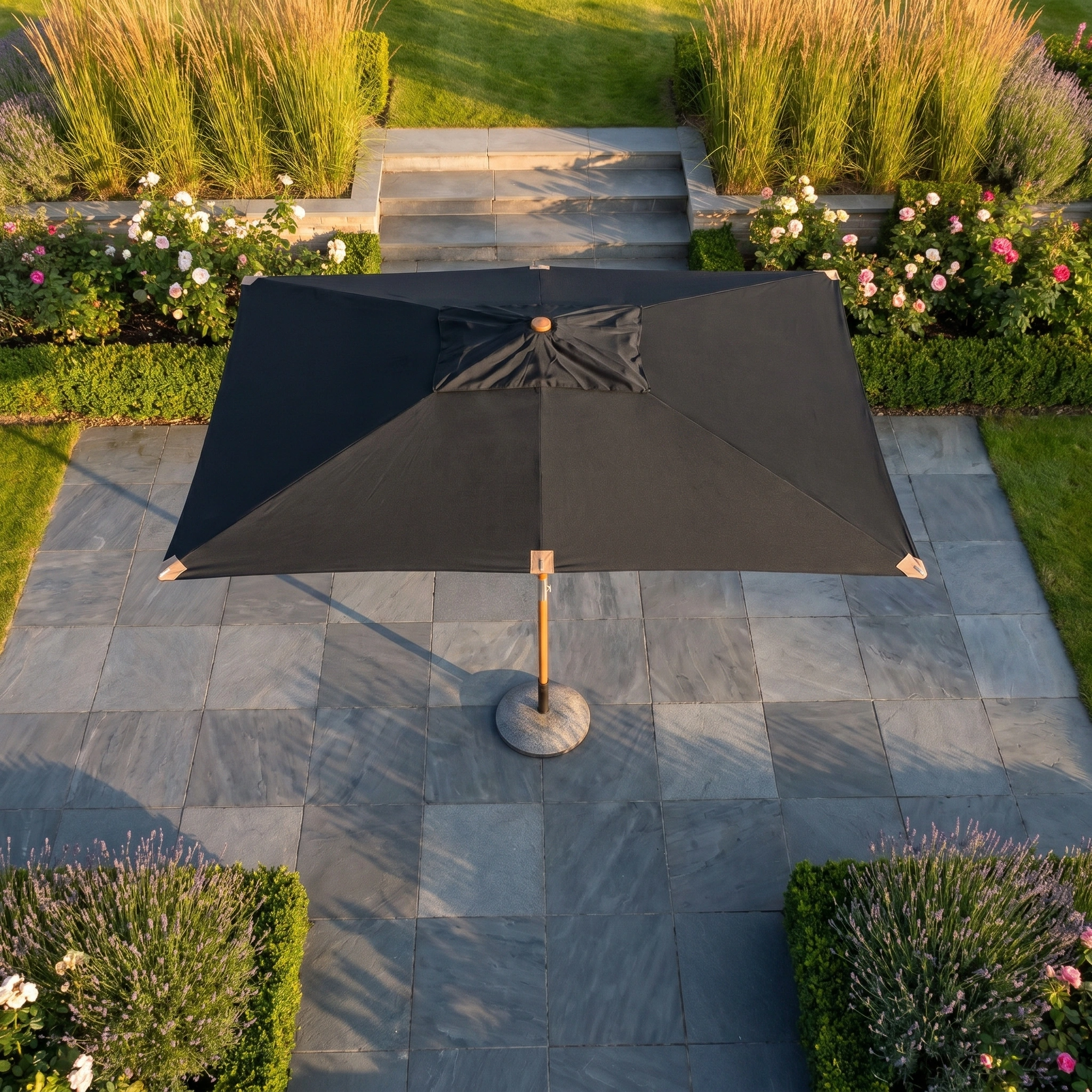 The Styx 3m x 2m Rectangular Deluxe Wooden Parasol in Black stands on a stone terrace, surrounded by manicured hedges and lush garden beds.