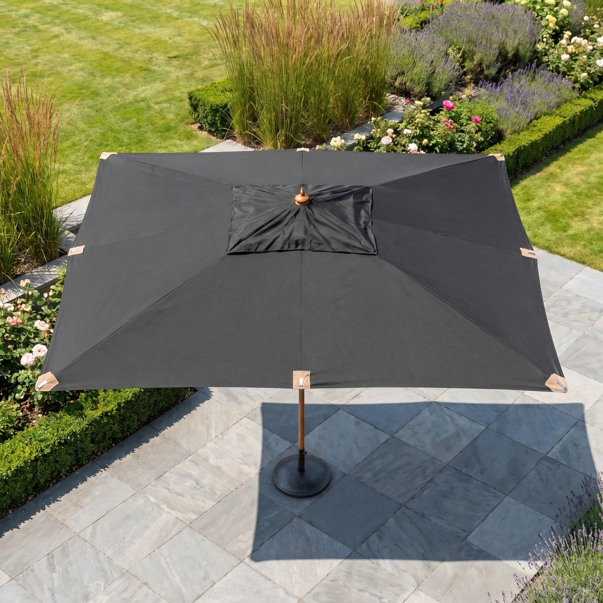 The Styx 3m x 2m Rectangular Deluxe Wooden Parasol in Black stands on a stone terrace, surrounded by green grass and vibrant garden plants.
