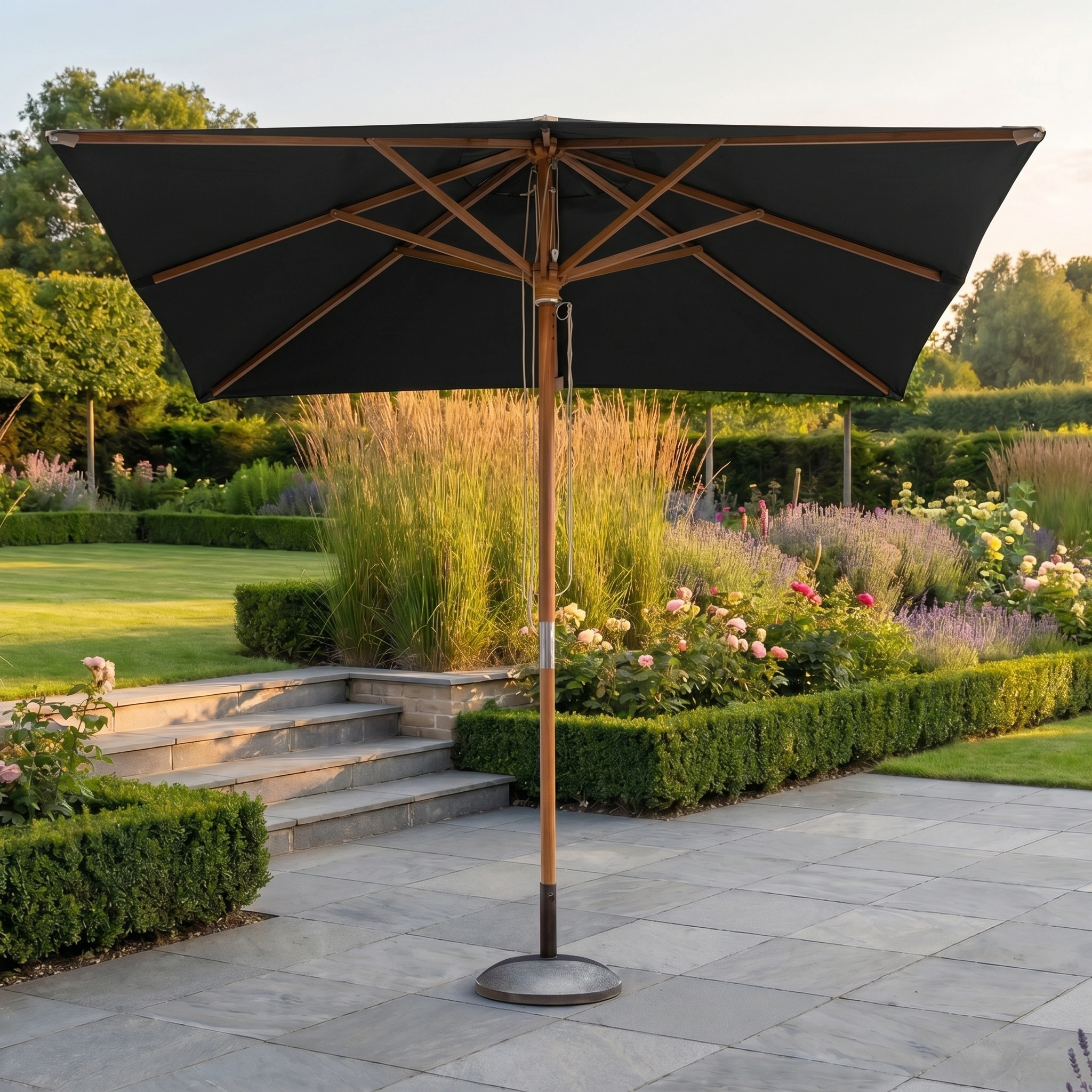 The Styx 3m x 2m Rectangular Deluxe Wooden Parasol in Black stands open on a stone terrace in a landscaped garden.