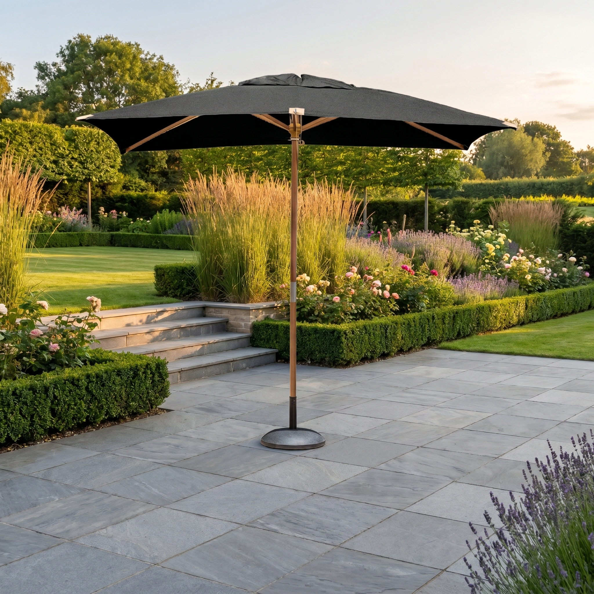 The Styx 3m x 2m Rectangular Deluxe Wooden Parasol in Black sits on a stone terrace, surrounded by vibrant flowers and trimmed hedges, creating the perfect garden retreat.