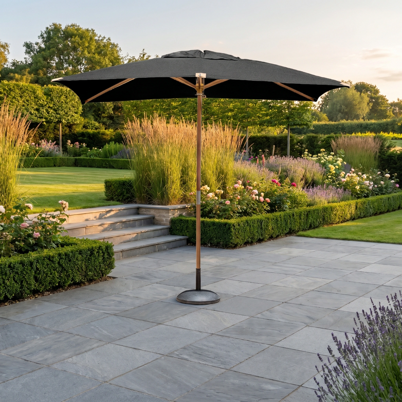 The Styx 3m x 2m Rectangular Deluxe Wooden Parasol in Black sits on a stone terrace, surrounded by vibrant flowers and trimmed hedges, creating the perfect garden retreat.