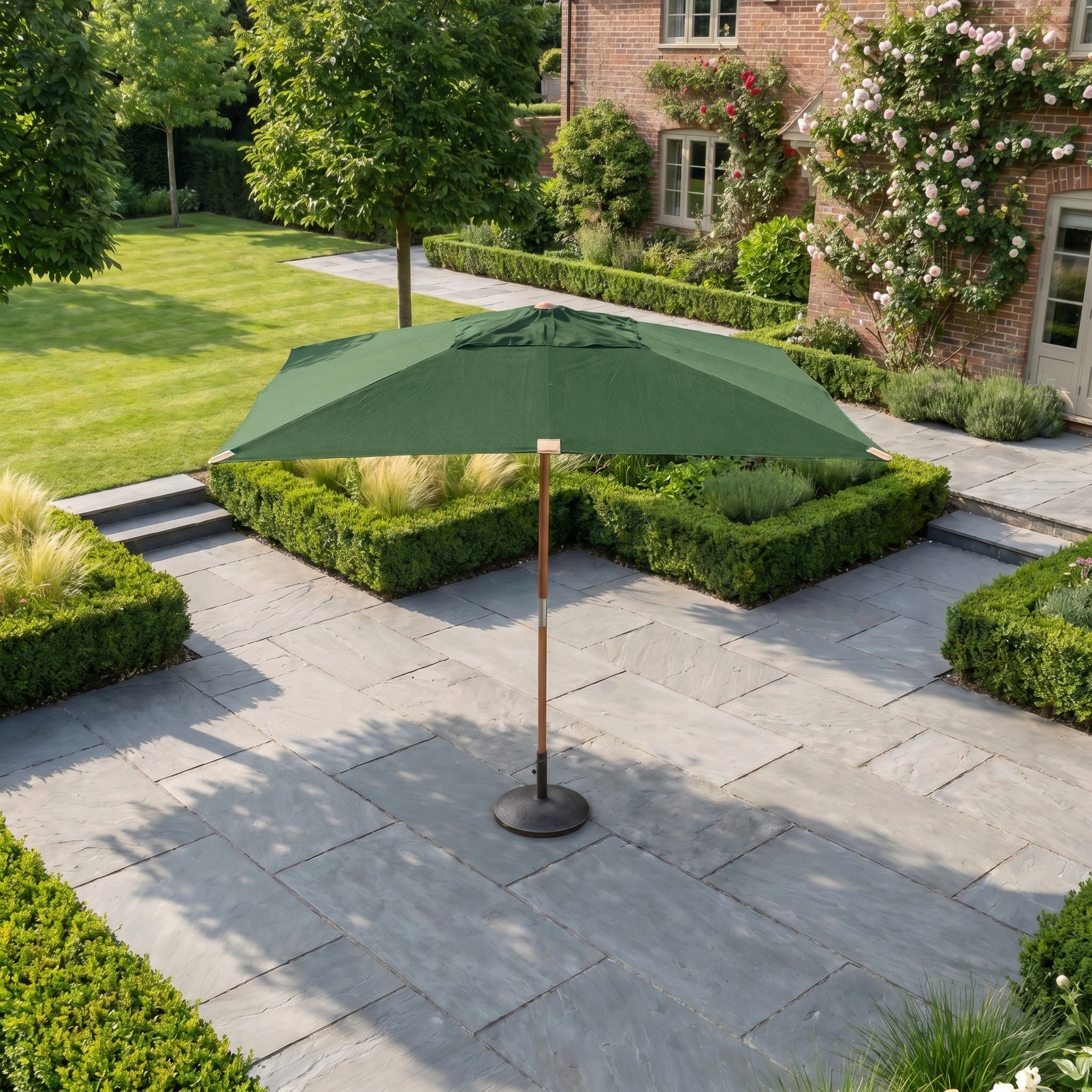 The Styx 3m x 2m Rectangular Deluxe Wooden Parasol in Green stands on a stone terrace, bordered by manicured hedges and a garden beside a brick house.