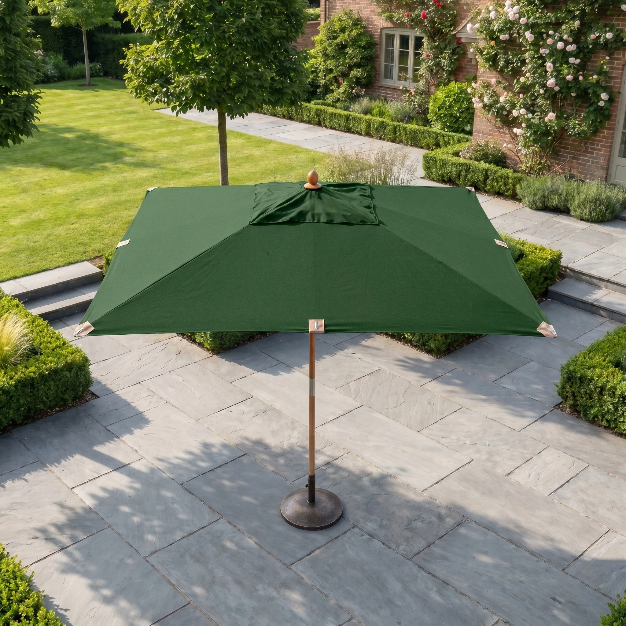 The Styx 3m x 2m Rectangular Deluxe Wooden Parasol in Green features a wooden pole and base, and sits on a stone patio surrounded by manicured gardens.
