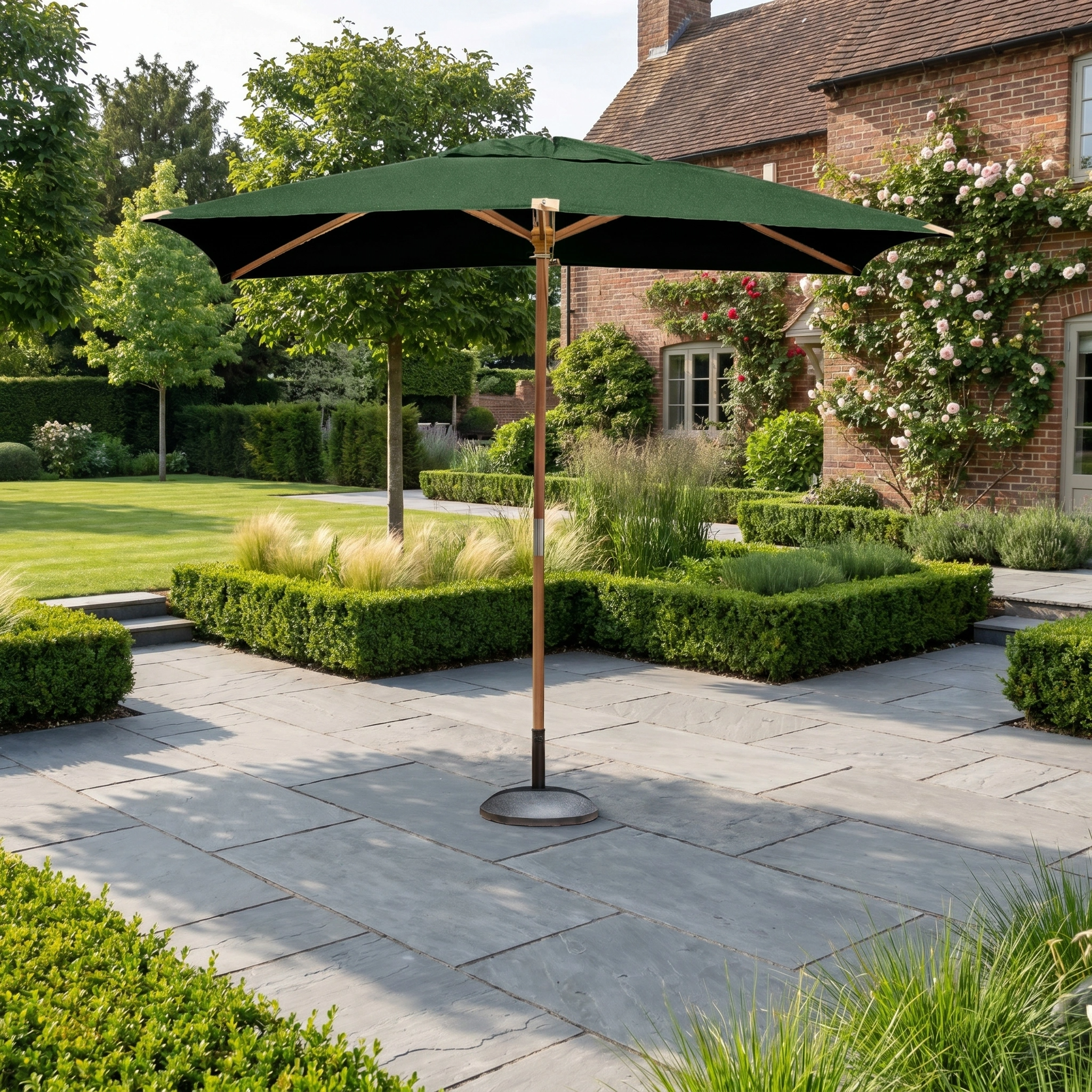 The Styx 3m x 2m Rectangular Deluxe Wooden Parasol in Green stands on a stone terrace in a landscaped garden by a brick house, providing elegant shade with its spacious canopy.