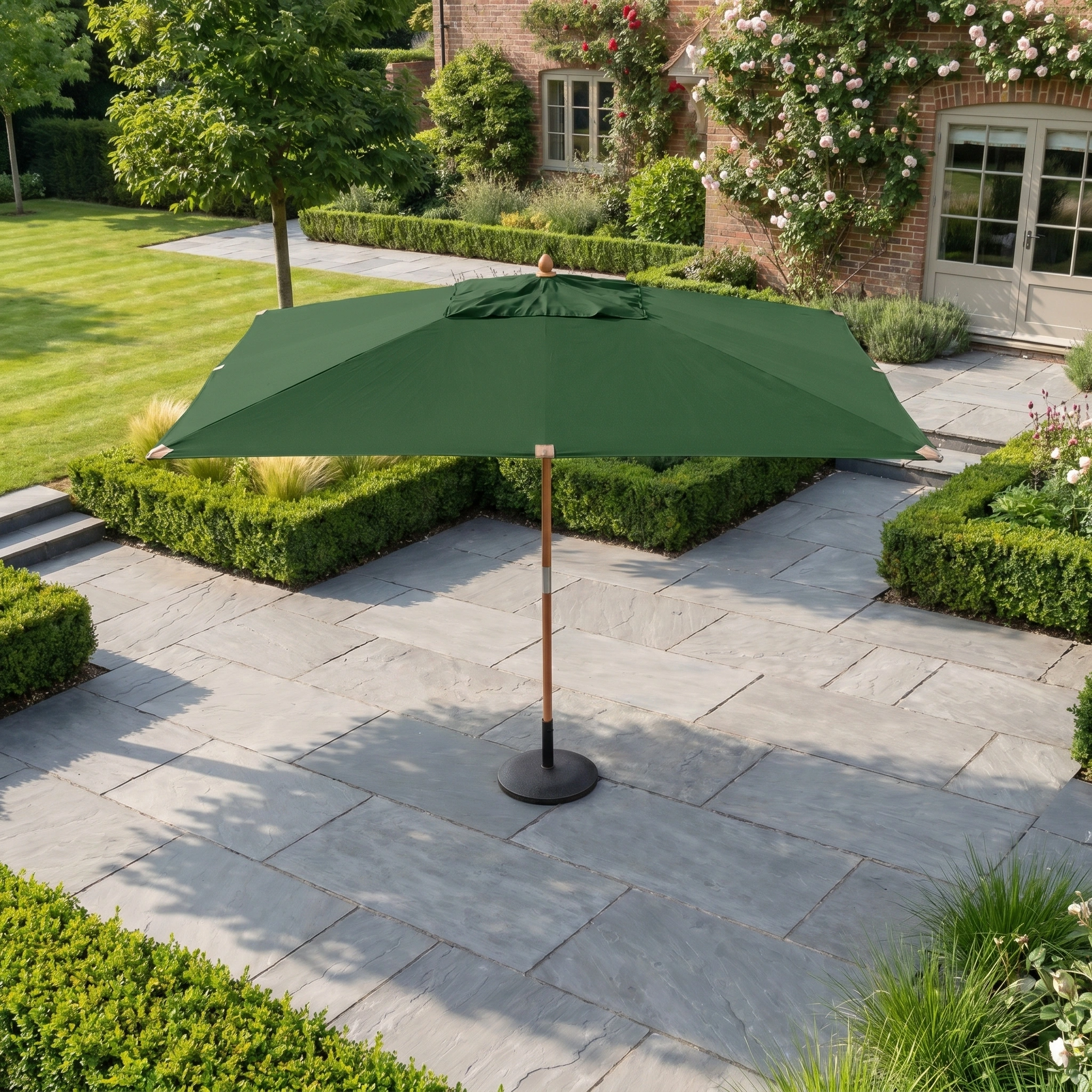 The Styx 3m x 2m Rectangular Deluxe Wooden Parasol in Green enhances a stone terrace, encircled by hedges and blooming plants, creating a tranquil outdoor retreat.