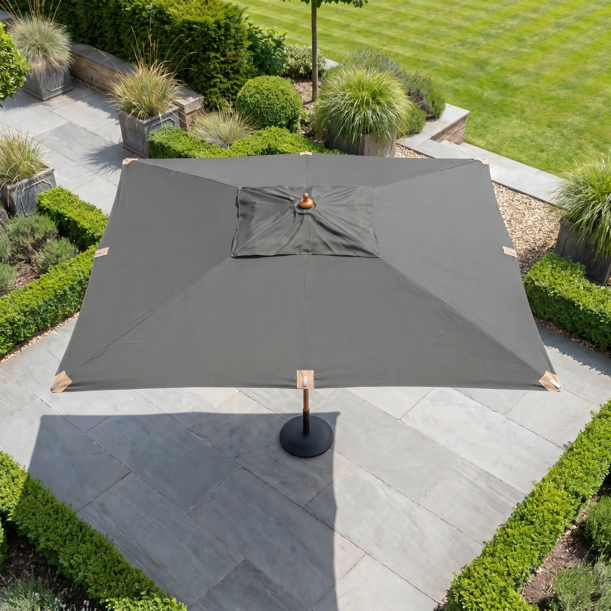 The Styx 3m x 2m Rectangular Deluxe Wooden Parasol in Grey stands on a stone terrace, bordered by green hedges and a landscaped garden.