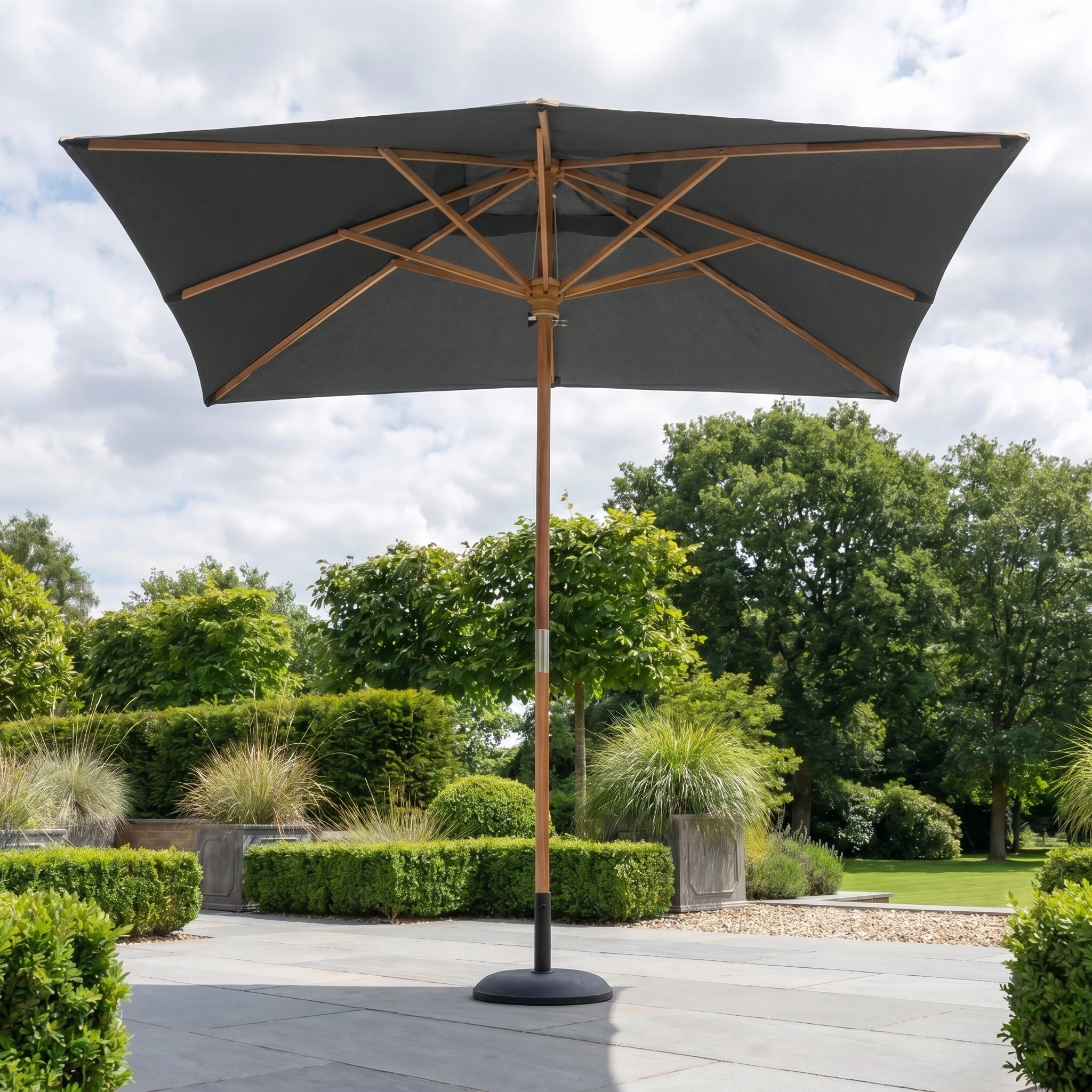 A Styx 3m x 2m Rectangular Deluxe Wooden Parasol in Grey stands on a stone terrace in a lush green garden beneath a cloudy sky.