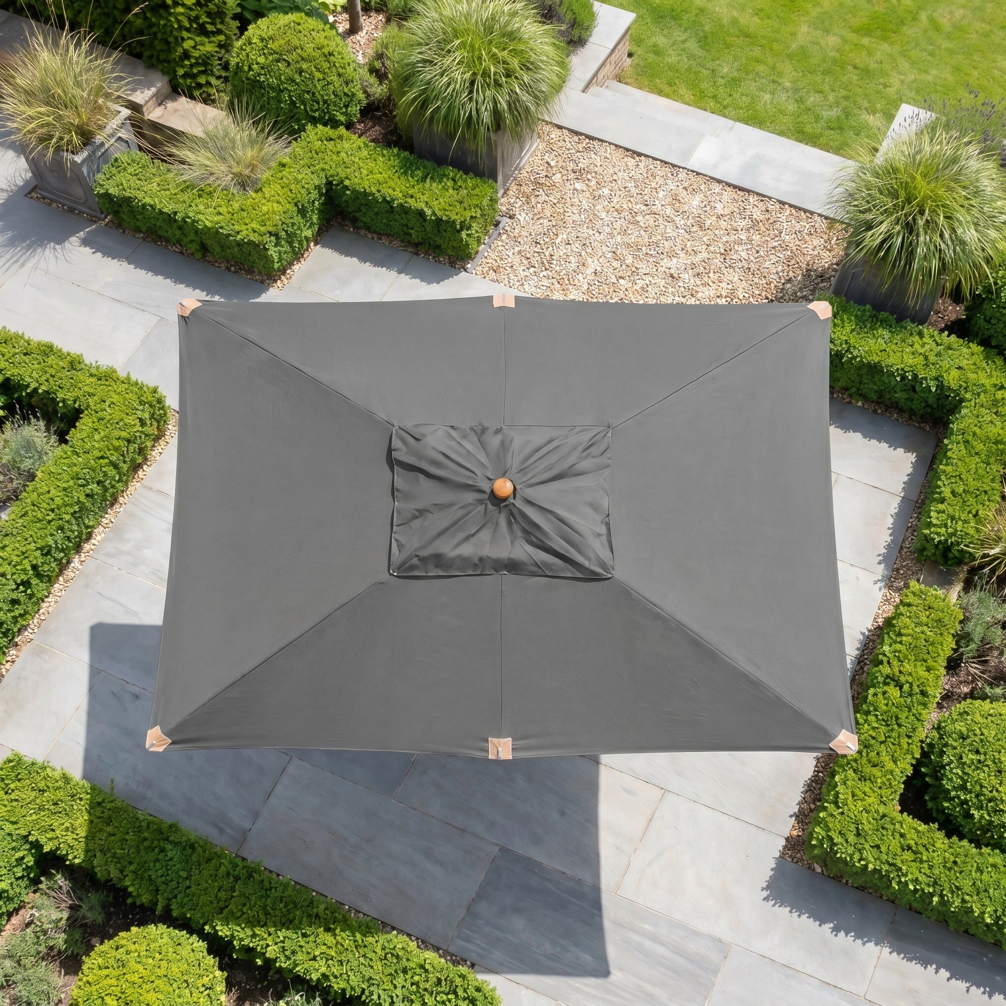 The Styx 3m x 2m Rectangular Deluxe Wooden Parasol in Grey shades stone tiles, bordered by neatly trimmed hedges and plants.