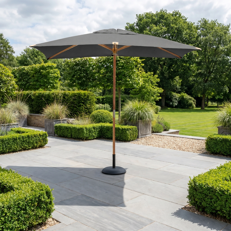 The Styx 3m x 2m Rectangular Deluxe Wooden Parasol in Grey stands on a stone terrace, surrounded by green shrubs and a lush lawn.