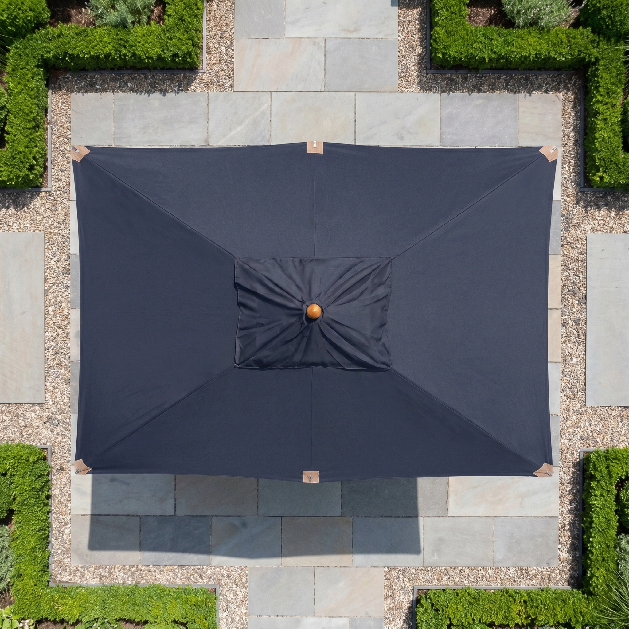 A Styx 3m x 2m Rectangular Deluxe Wooden Parasol in Navy is centered on a stone patio bordered by green hedges, viewed from above.