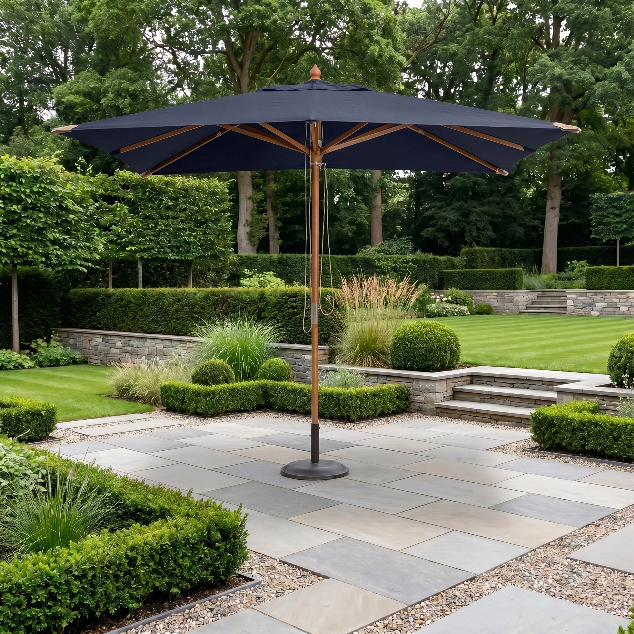 The Styx 3m x 2m Rectangular Deluxe Wooden Parasol in Navy is set in a landscaped garden featuring stone paving and trimmed greenery.