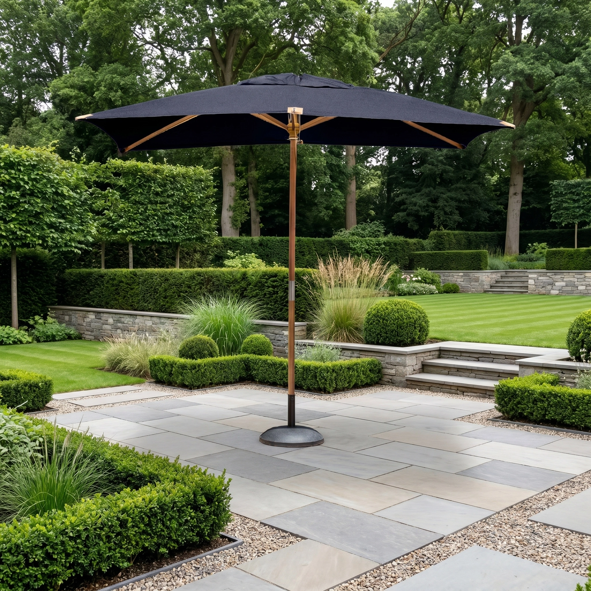 The Styx 3m x 2m Rectangular Deluxe Wooden Parasol in Navy stands on a stone terrace in a landscaped garden with trimmed hedges and trees.