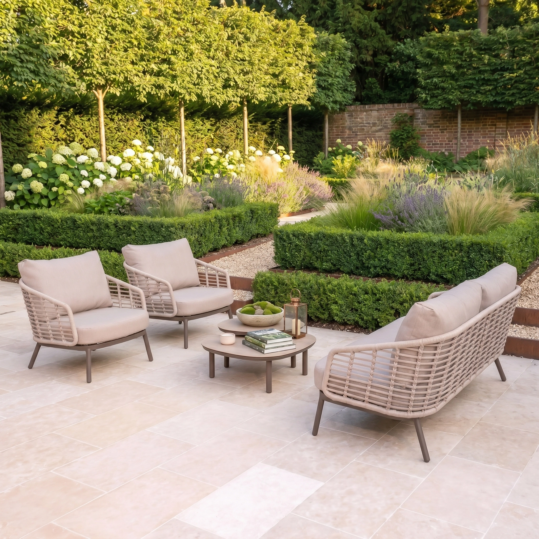 The Sutera 3 Seat Sofa Set in Putty sits on a stone patio with all-weather cushions, framed by a lush, manicured garden backdrop.