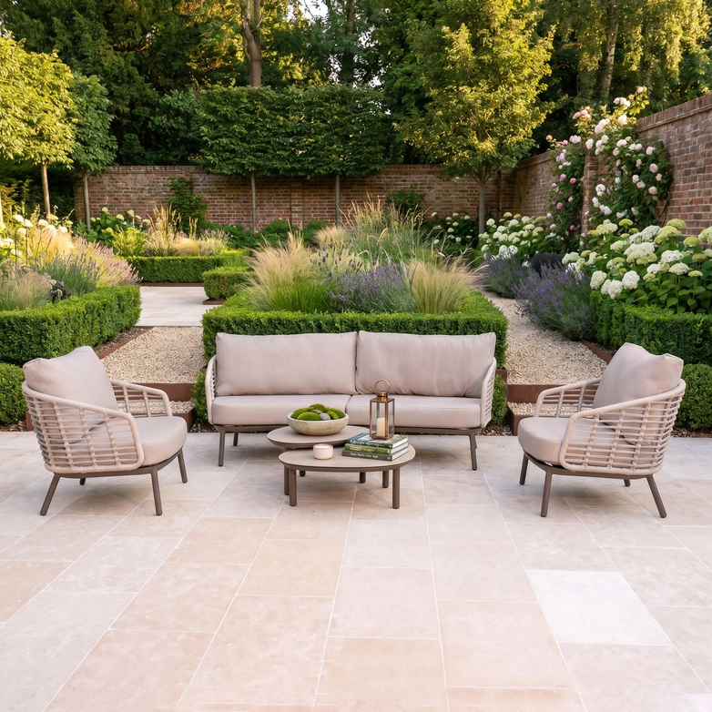Outdoor patio with the Sutera 3 Seat Sofa Set in Putty, featuring beige all-weather cushions and coffee tables, set amidst lush greenery for a stylish garden furniture ensemble.